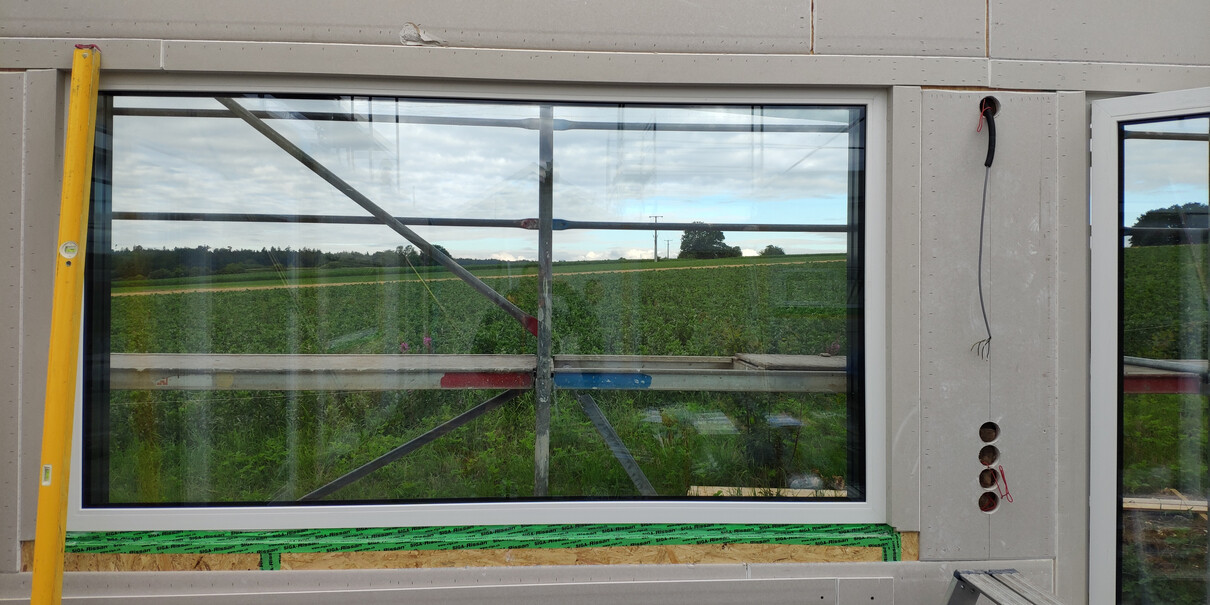 Außenwand mit großem Fenster, Schalung und Gerüst auf Baustelle, Blick ins Grüne.