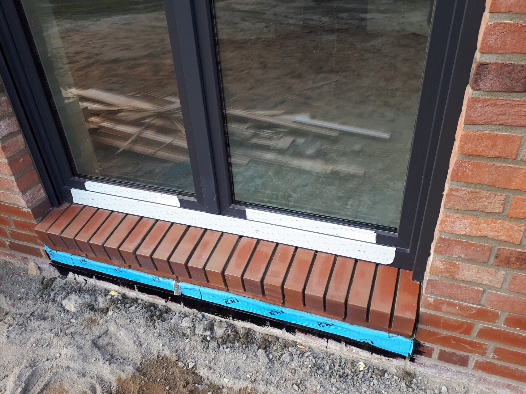 Schwarzes Metallfenster in roter Backsteinwand; Bauarbeiten, blaues Band unten, Holzlatten hinter Glas.