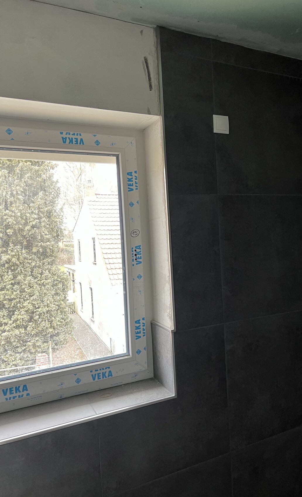 Neues Fenster mit VEKA-Schutzfolie in dunkler Fliesenwand; Blick nach außen auf Bäume und Dächer.