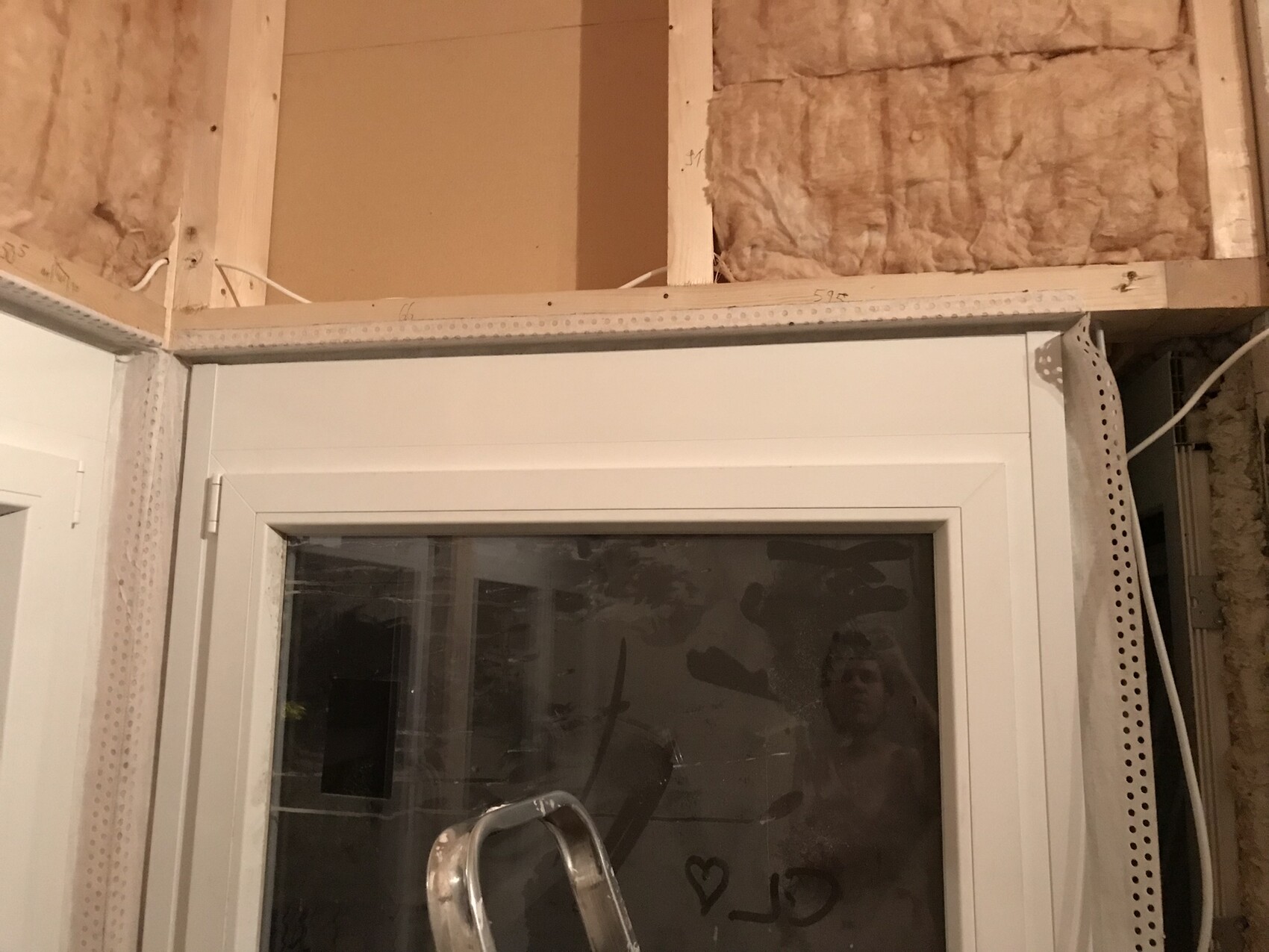 Unverkleidete Bauwand mit Dämmung, weißem Fensterrahmen; Kabel, Spiegelung einer Person im Glas.
