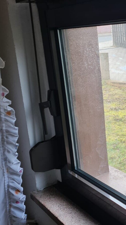 Innenfenster mit schwarzem Griff, Vorhang links, Blick nach außen auf braune Wand und Grünfläche.