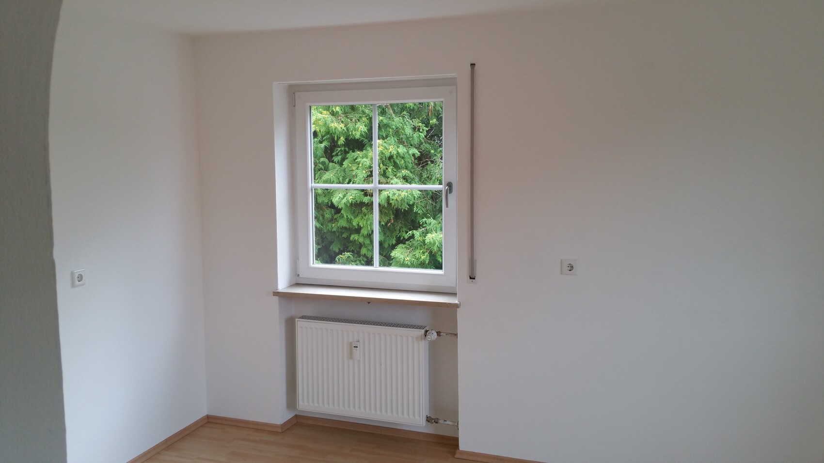 Kleines Zimmer mit weißem Fenster, Heizkörper darunter, Blick auf grüne Bäume