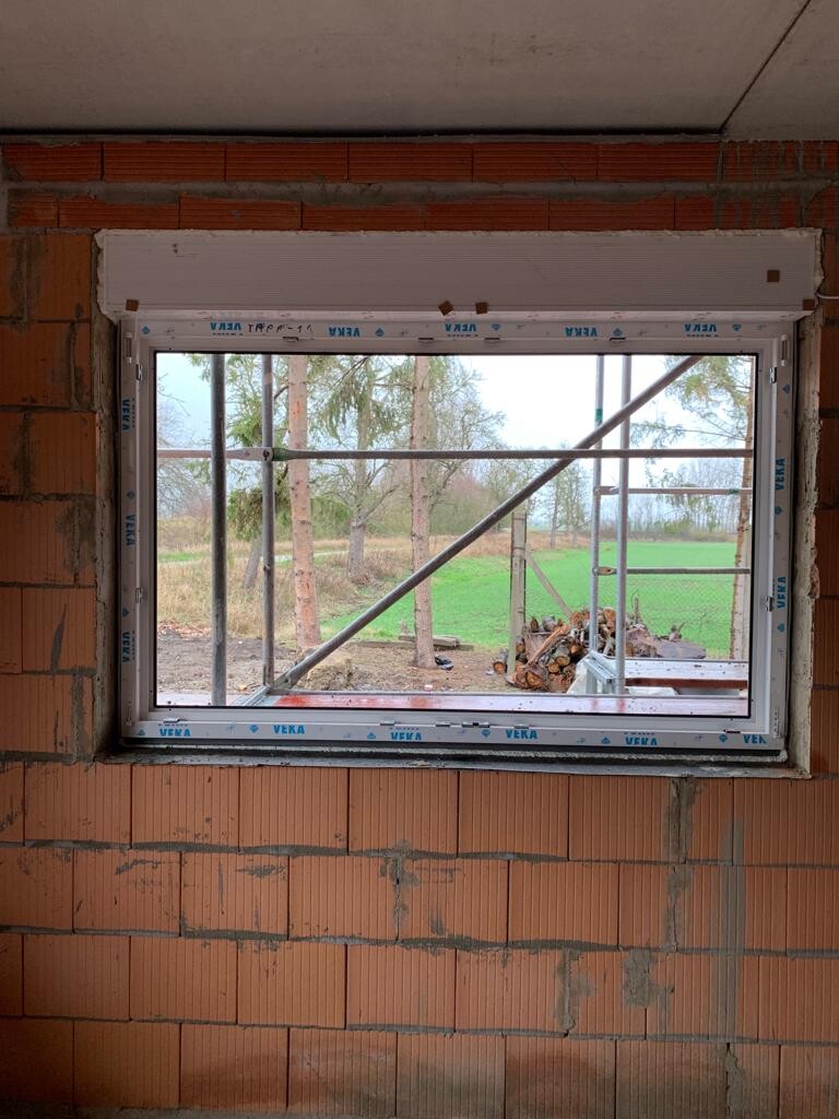 Fenster in roter Ziegelwand unter Baugerüst; Blick nach Außen auf Bäume und Wiese.