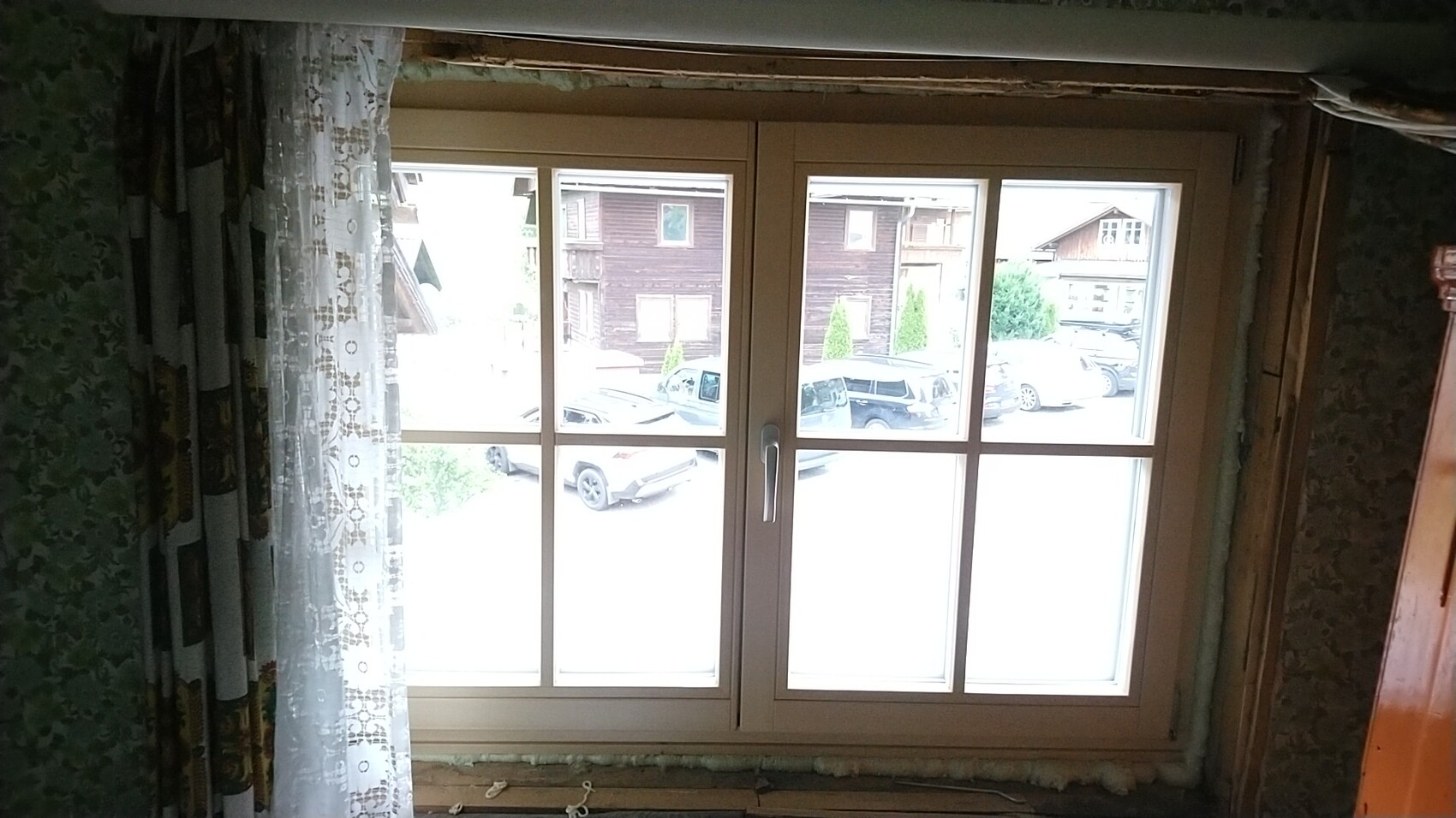 Fenster mit hellem Rahmen, blick nach draußen auf geparkte Autos vor Holzhäusern; linke Spitze-Vorhänge.