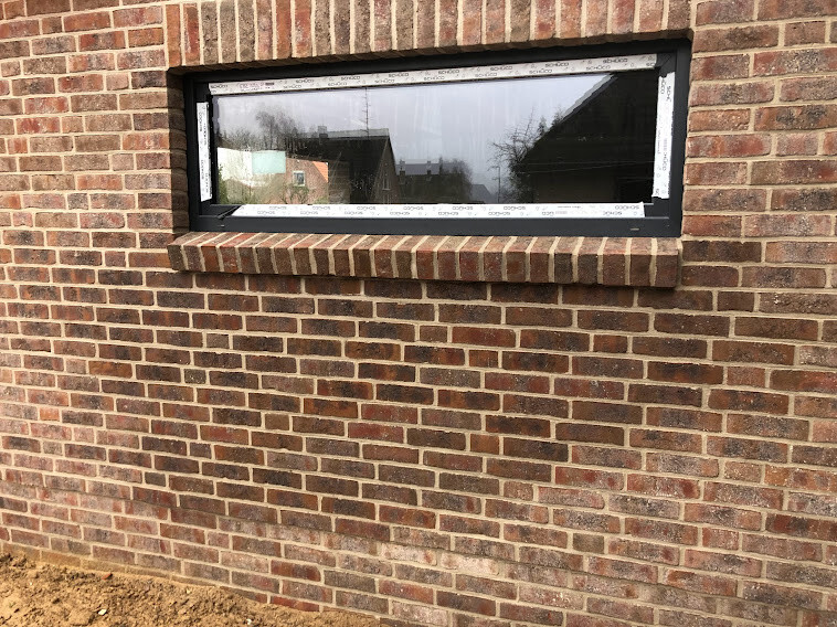 Kleines, rechteckiges Fenster in roter Backsteinwand; Glas spiegelt den Außenbereich.