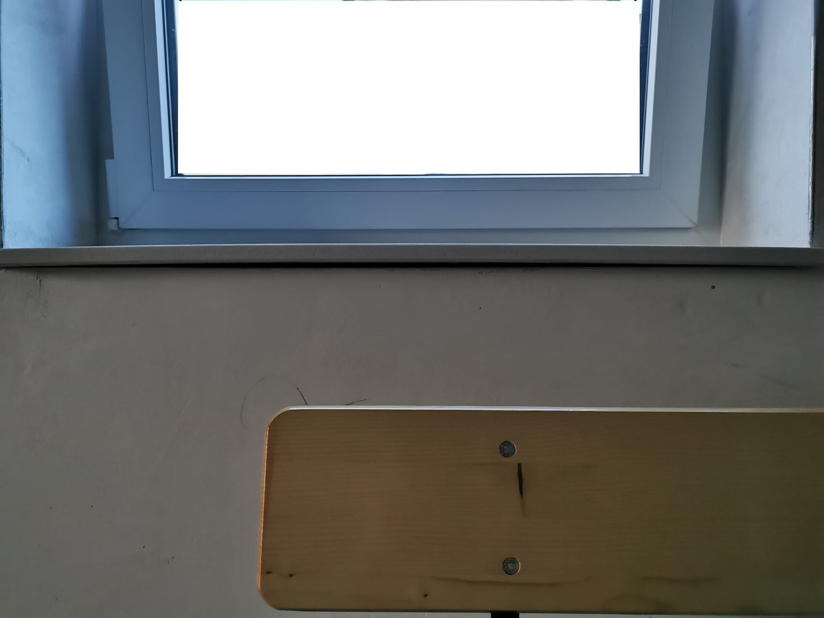 Innenraum: Holzbank unten rechts vor grauer Wand, großes Fenster oben.
