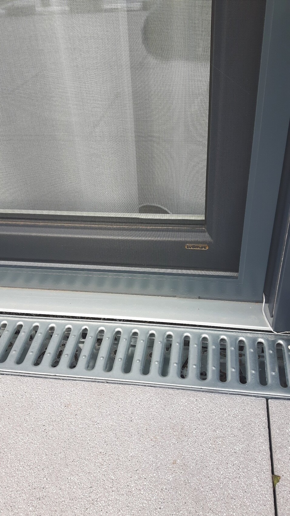 Fenster mit Fliegengitter und Metallrahmen; unten eine graue Bodenleiste mit Lüftungsschlitzen.