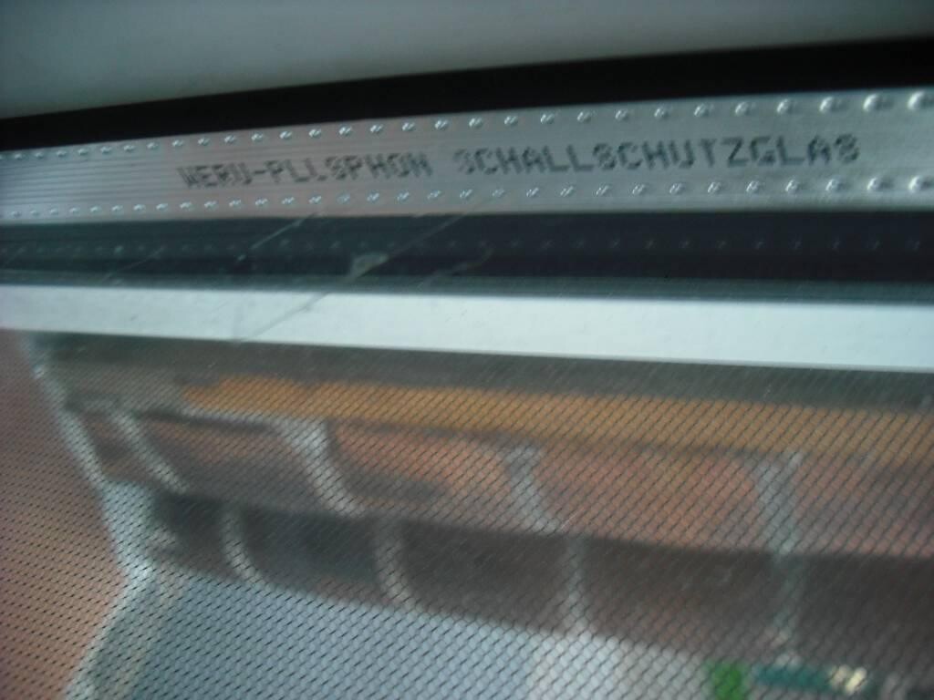 Blick durch ein Fenster mit Fliegengitter; oben eine gelochte Metallleiste mit eingraviertem Text.