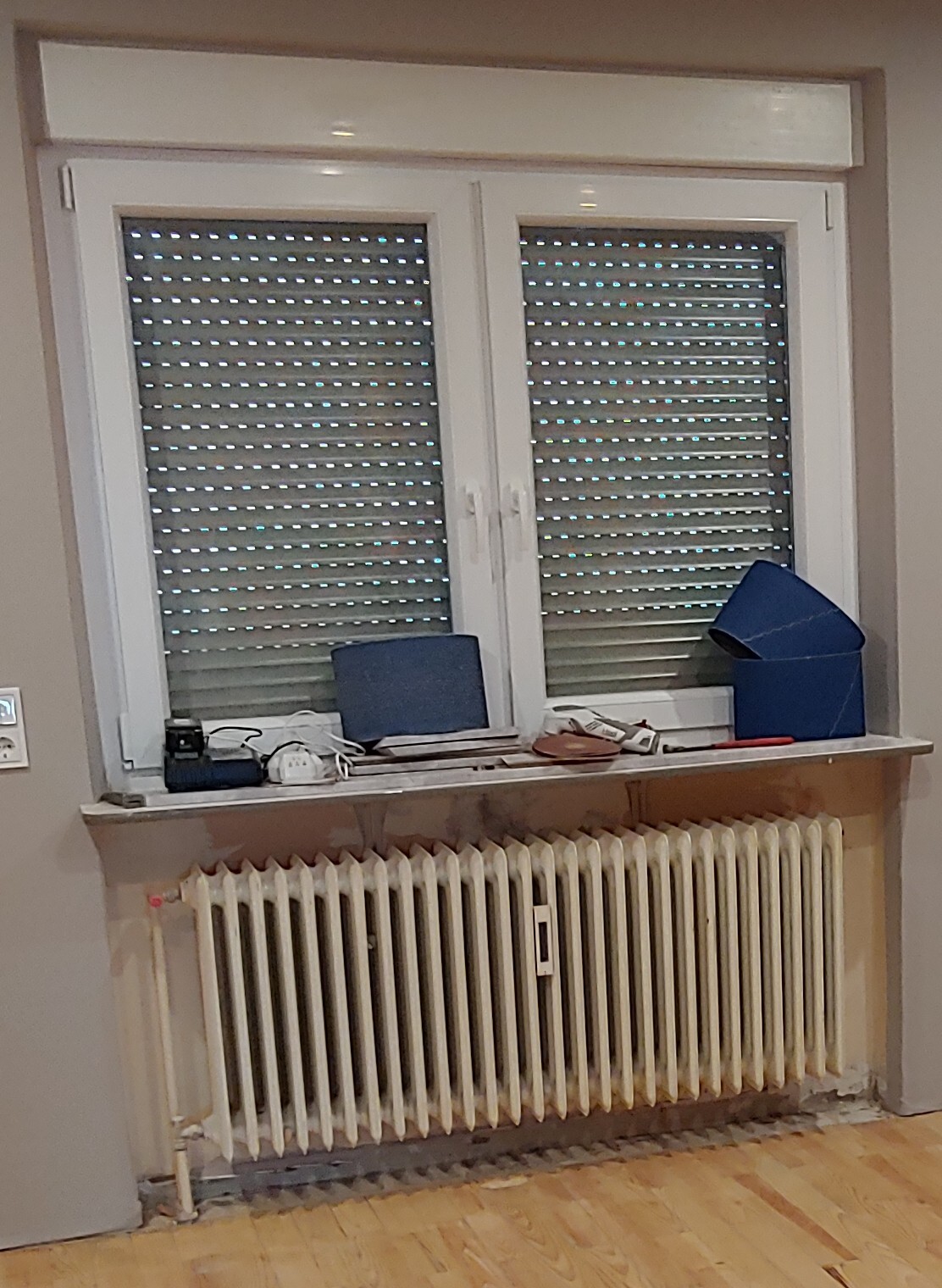 Fenster mit Rollläden und Heizkörper darunter; Fensterbank mit blauem Ordner, Box und Kabeln.