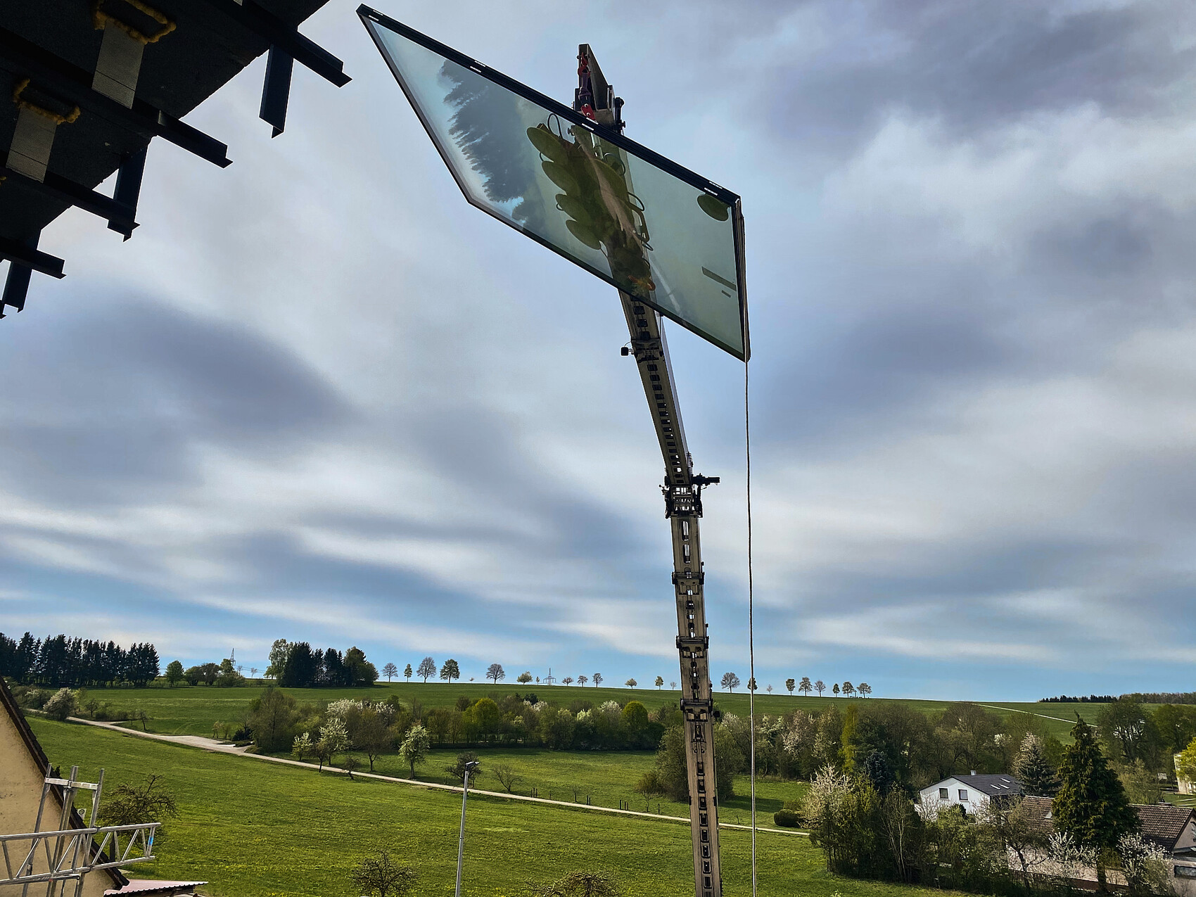 Kran hebt großes Glasscheibenpaneel an Gebäudekante; ländliche Landschaft im Hintergrund.