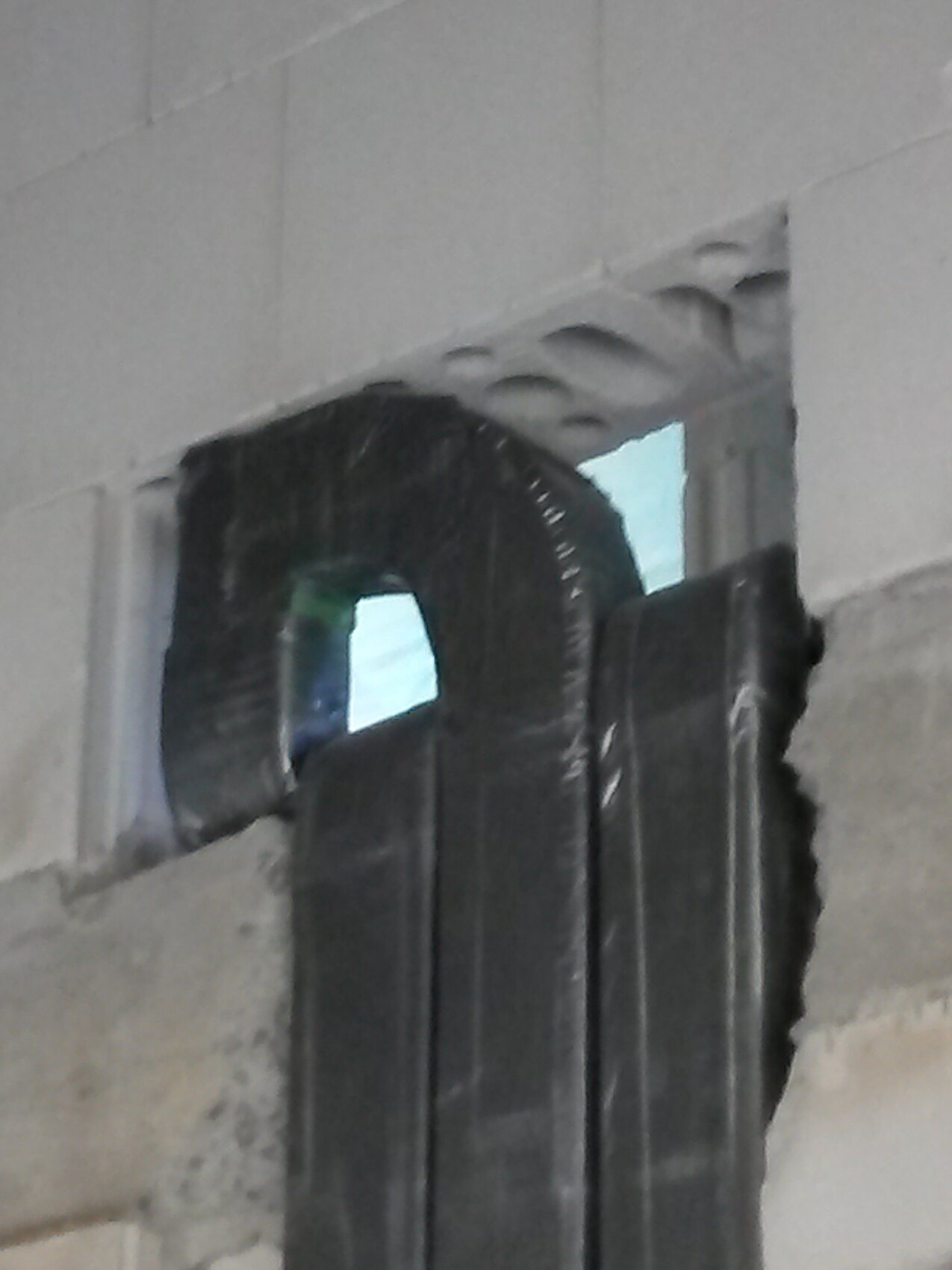 Große schwarze Rundform ragt durch eine Öffnung in einer grauen Bauwand; Bauplatten davor.