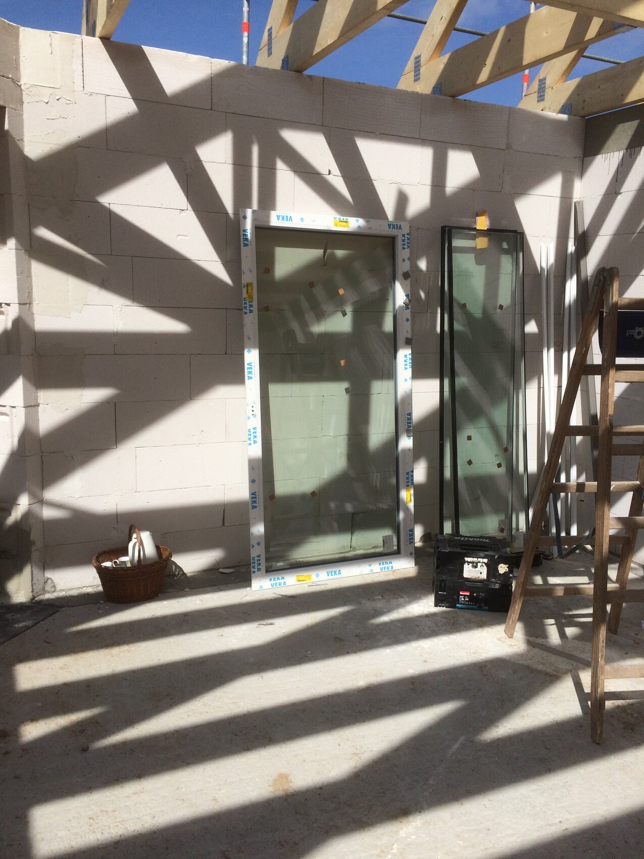 Baustelle mit zwei Fensterrahmen, Leiter, Werkzeugkasten und Korb; Schatten der Holzbalken.