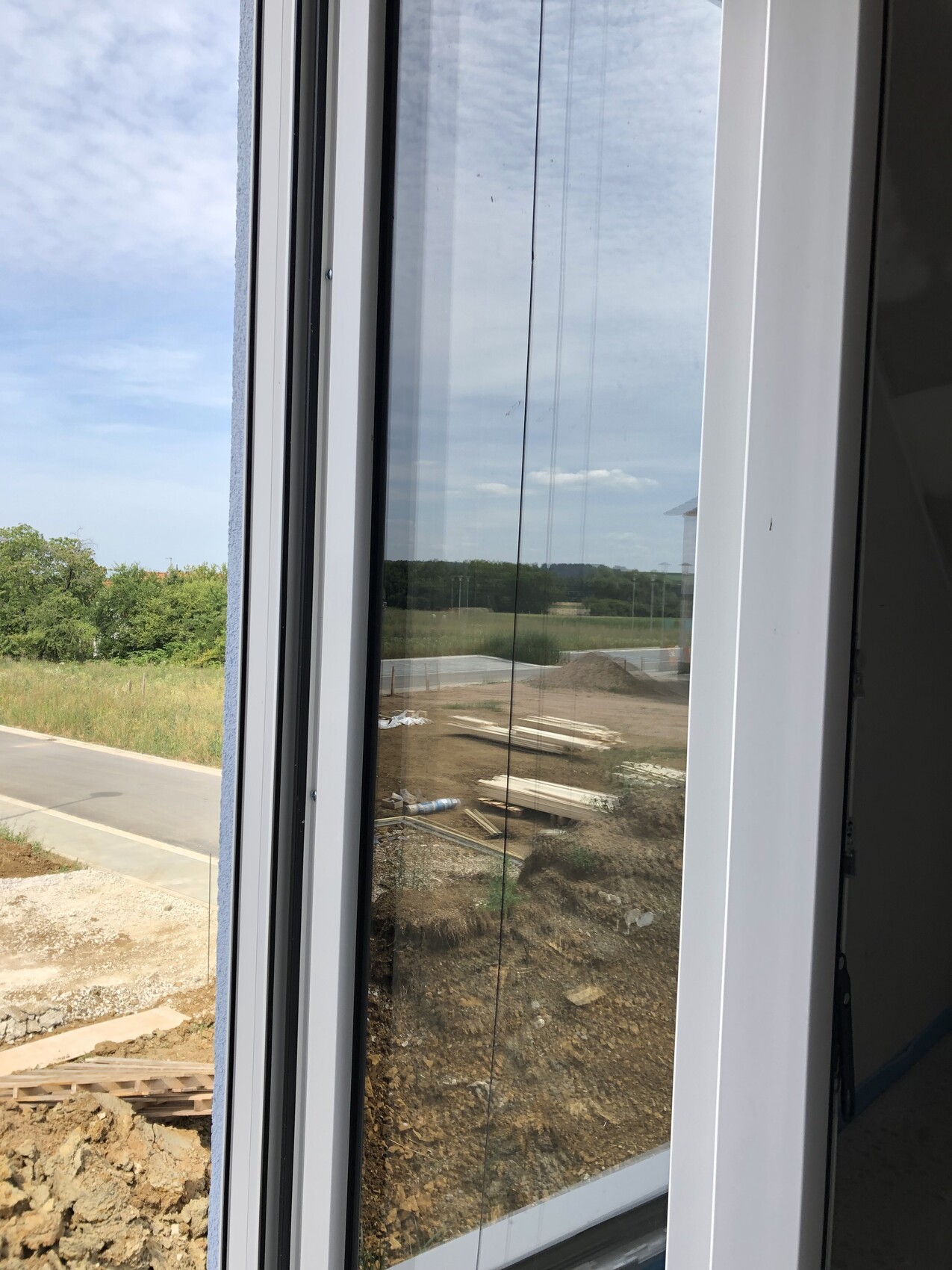 Blick aus Fenster auf Baustelle: Erde, Bretterstapel, Bauholz; Hintergrund Wald und Himmel.