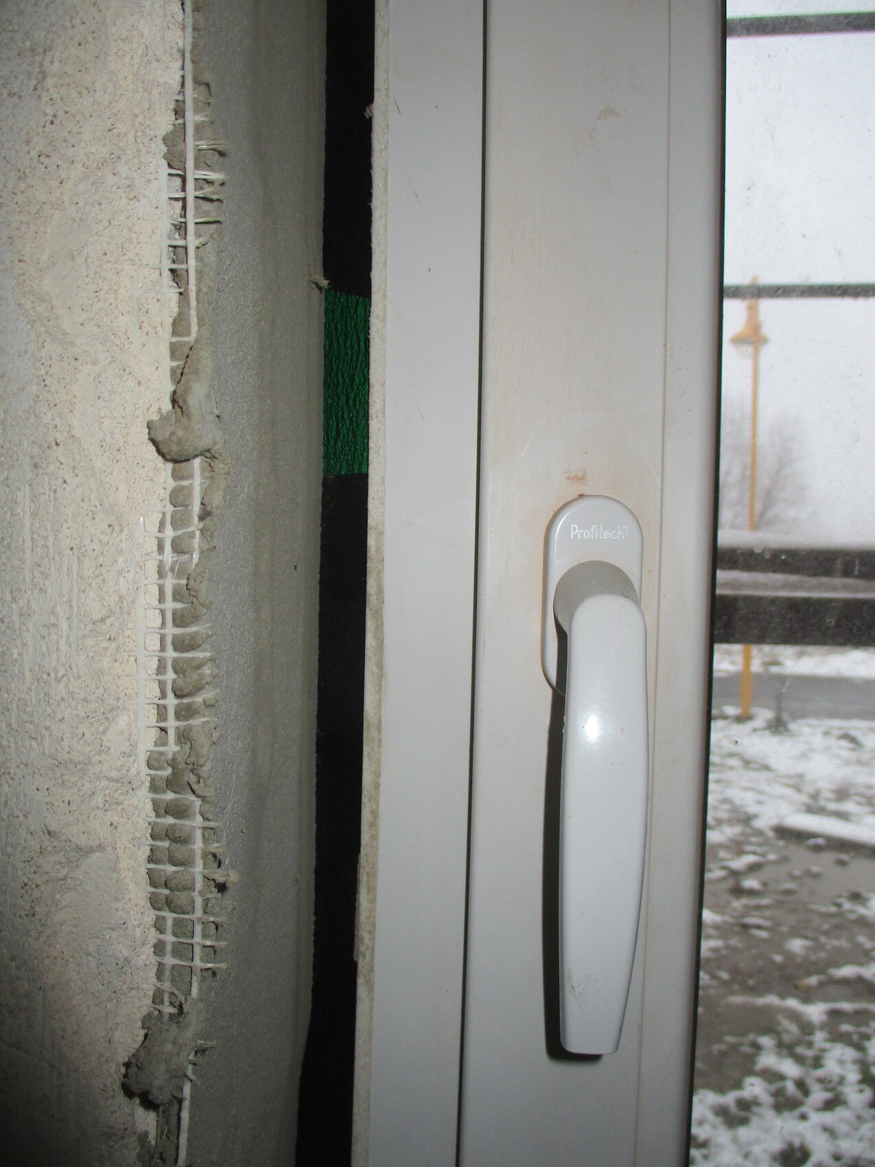 Nahaufnahme eines weißen Fenstergriffs am Türrahmen; daneben Wand mit Putzbruch.
