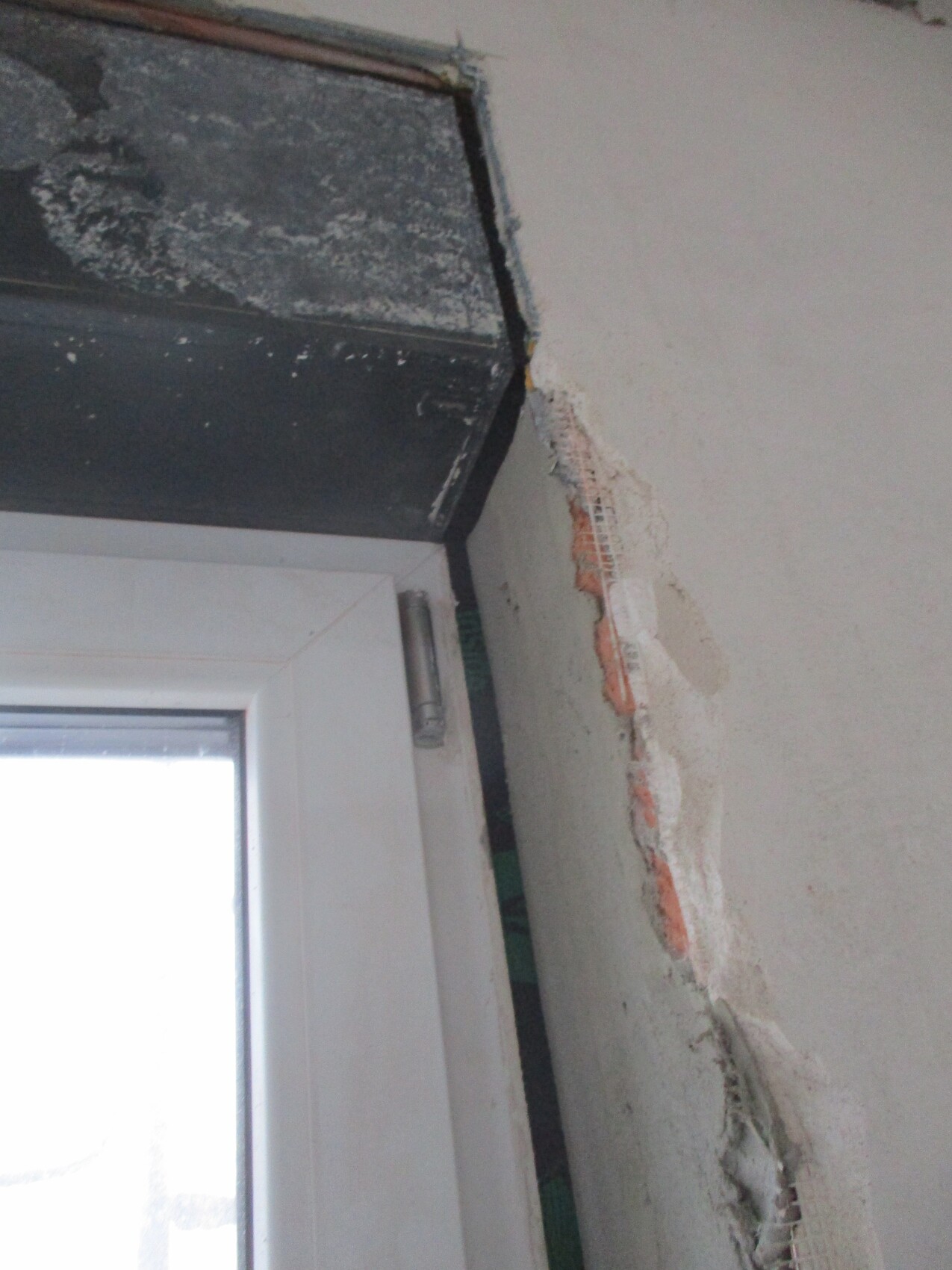 Fensterrahmen neben beschädigter Wand; freiliegter Ziegel, Putzreste und Drahtgewebe sichtbar.