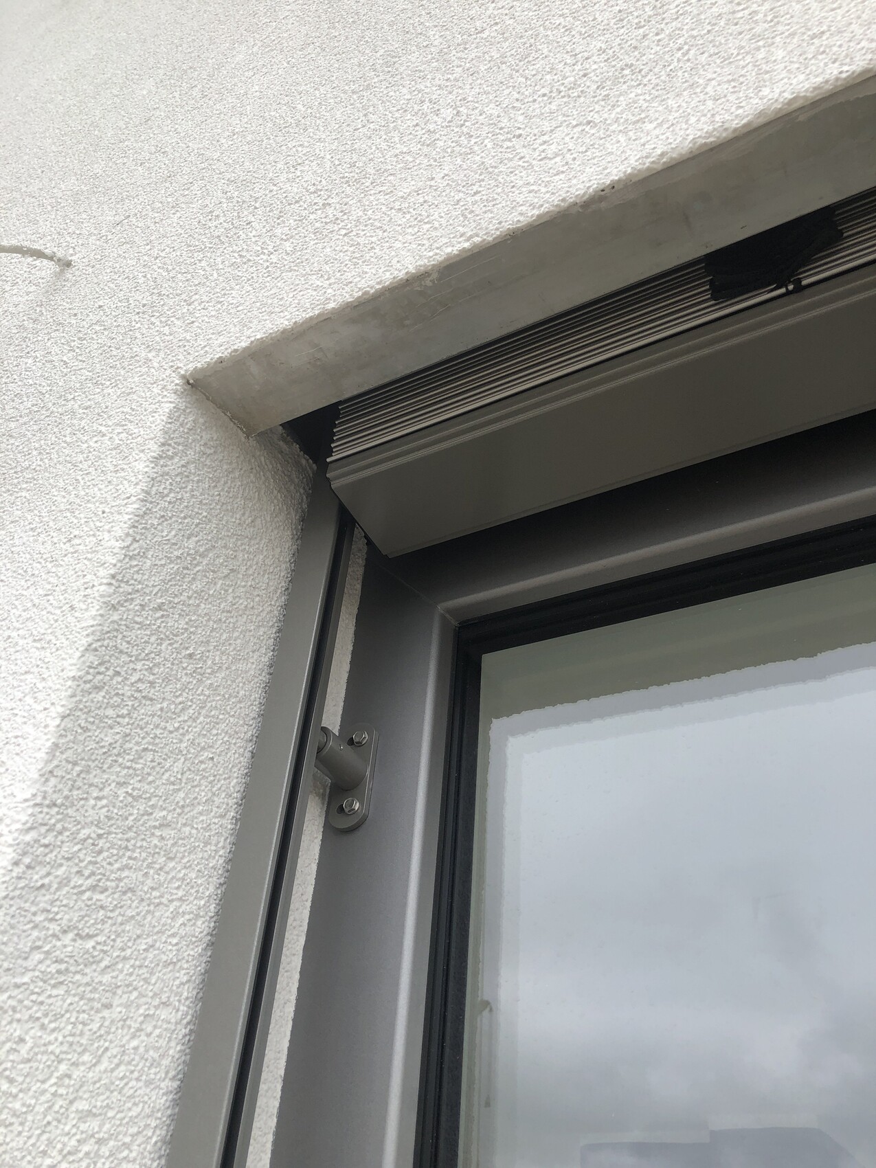 Grauer Fensterrahmen mit Rollladenführung über einer grob strukturierten Wand
