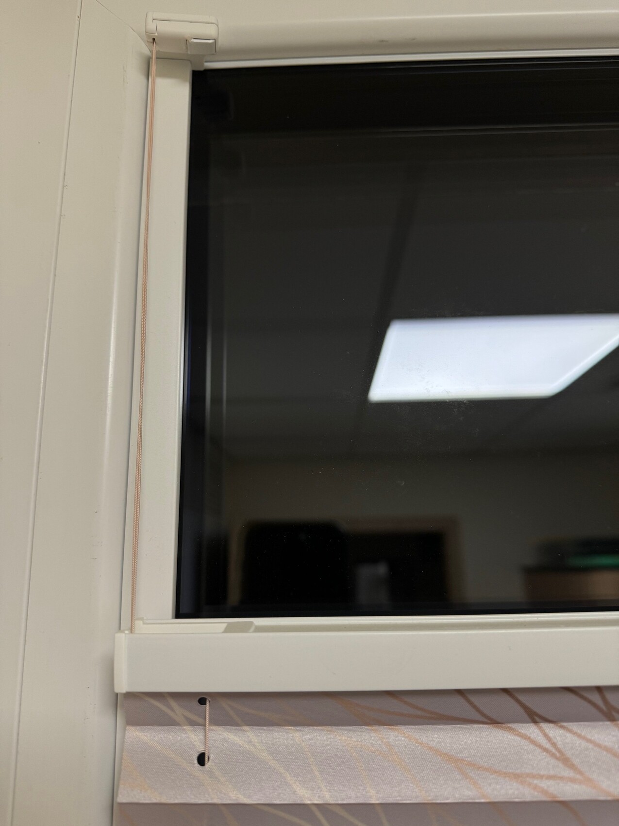 Weißer Fensterrahmen, linke Zugkette, dunkles Glas; unten gemustertes Stoffband sichtbar.