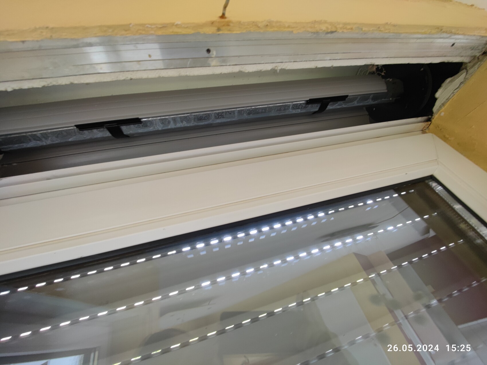 Nahaufnahme eines Fensterrahmens mit Rollladenführung und Glasfenster, gelb gestrichene Wand im Hintergrund