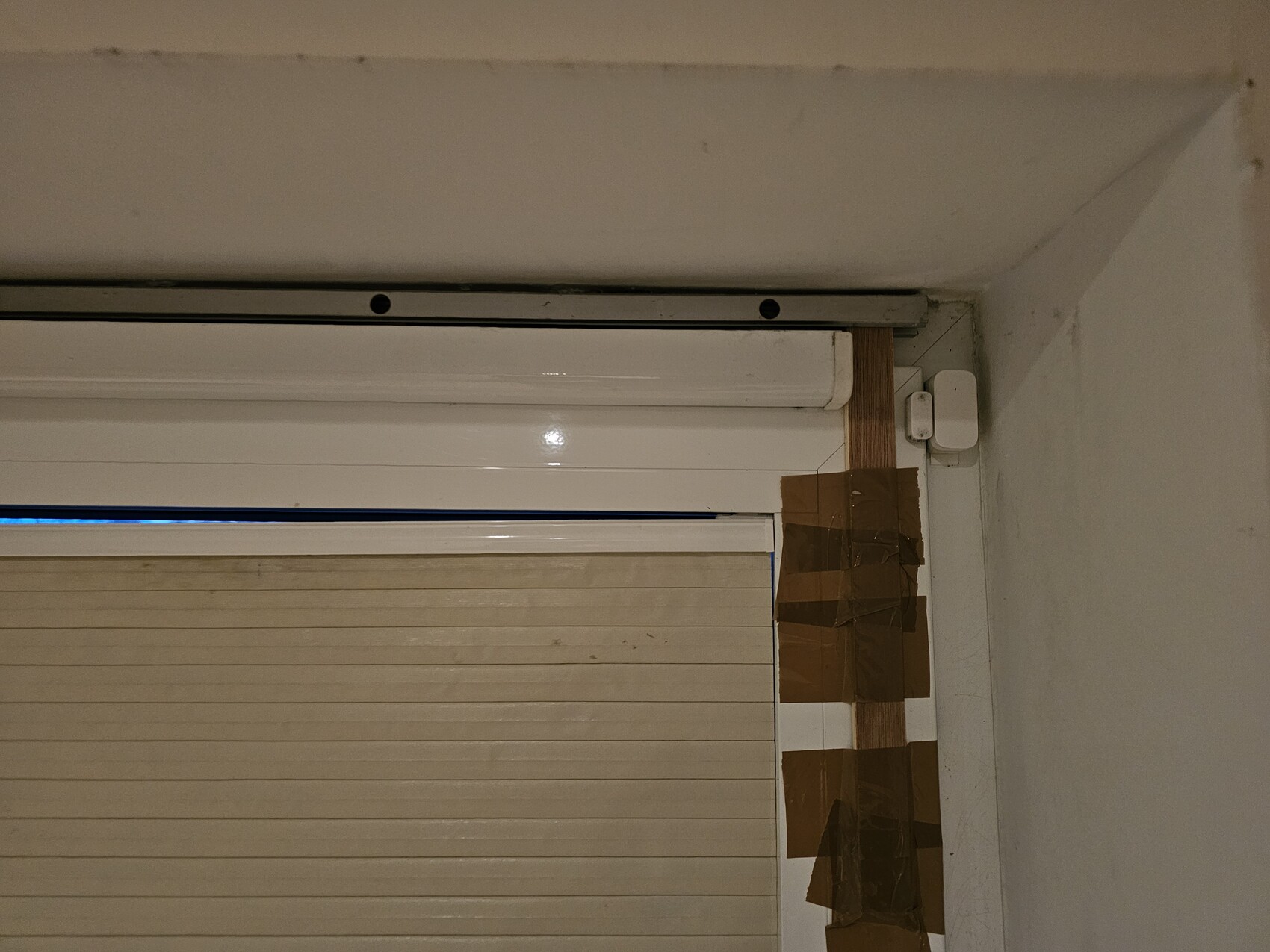 Beige horizontale Jalousie vor Fenster, oben Führungen, rechts Holzlatten mit braunem Klebeband.