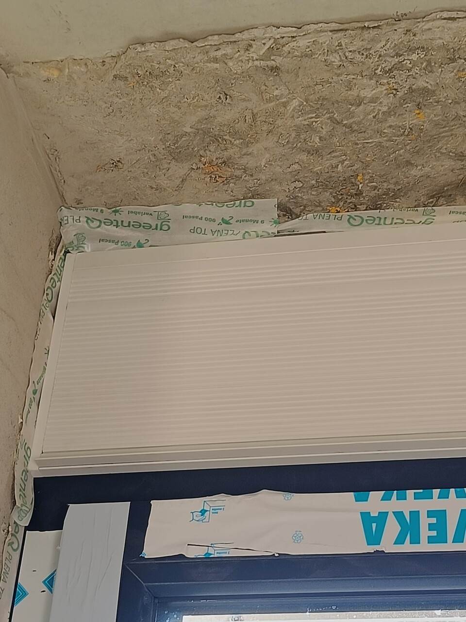 Weißer Rollladenkasten vor rauer Wand über blauem Türrahmen; Schutzfolie klebt am Rand.