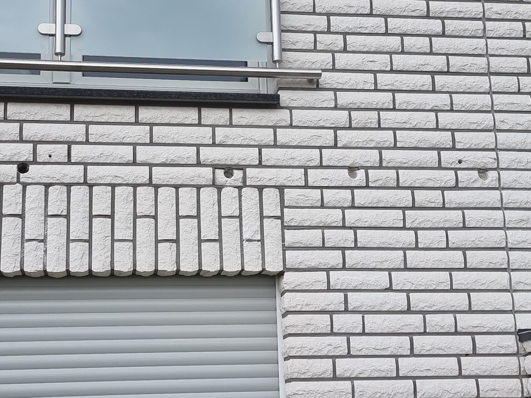 Weiße Ziegelwand mit Fenster, Metallgeländer und darunter Rollladen; Wand mit Löchern.