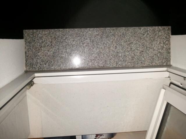 Fensterrahmen mit dunklem Granitsims an einer weißen Wand, offenes Türblatt sichtbar.