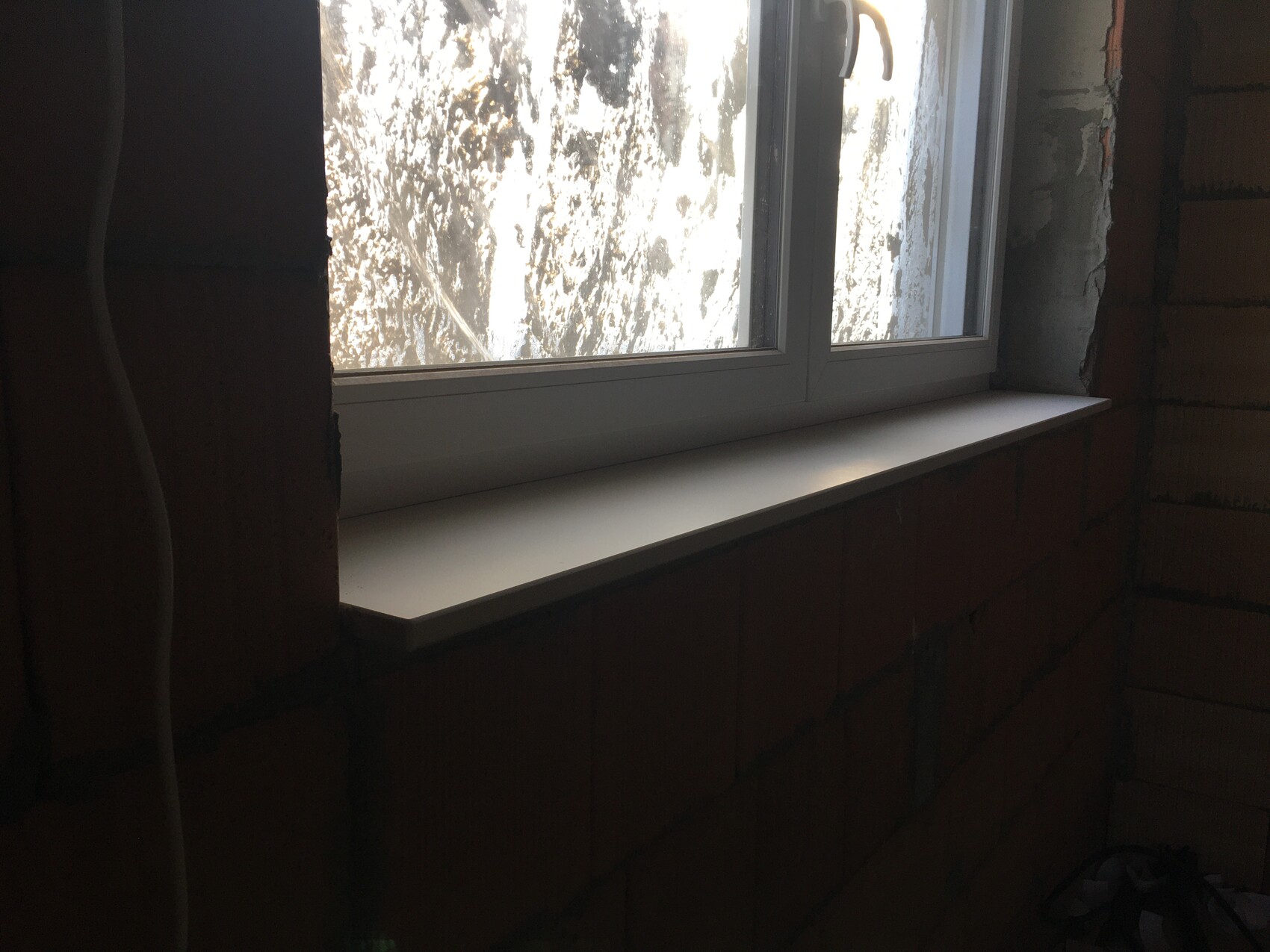 Fenster mit weißer Fensterbank in unfertiger Ziegelwand; draußen heller Lichtschein zu sehen.