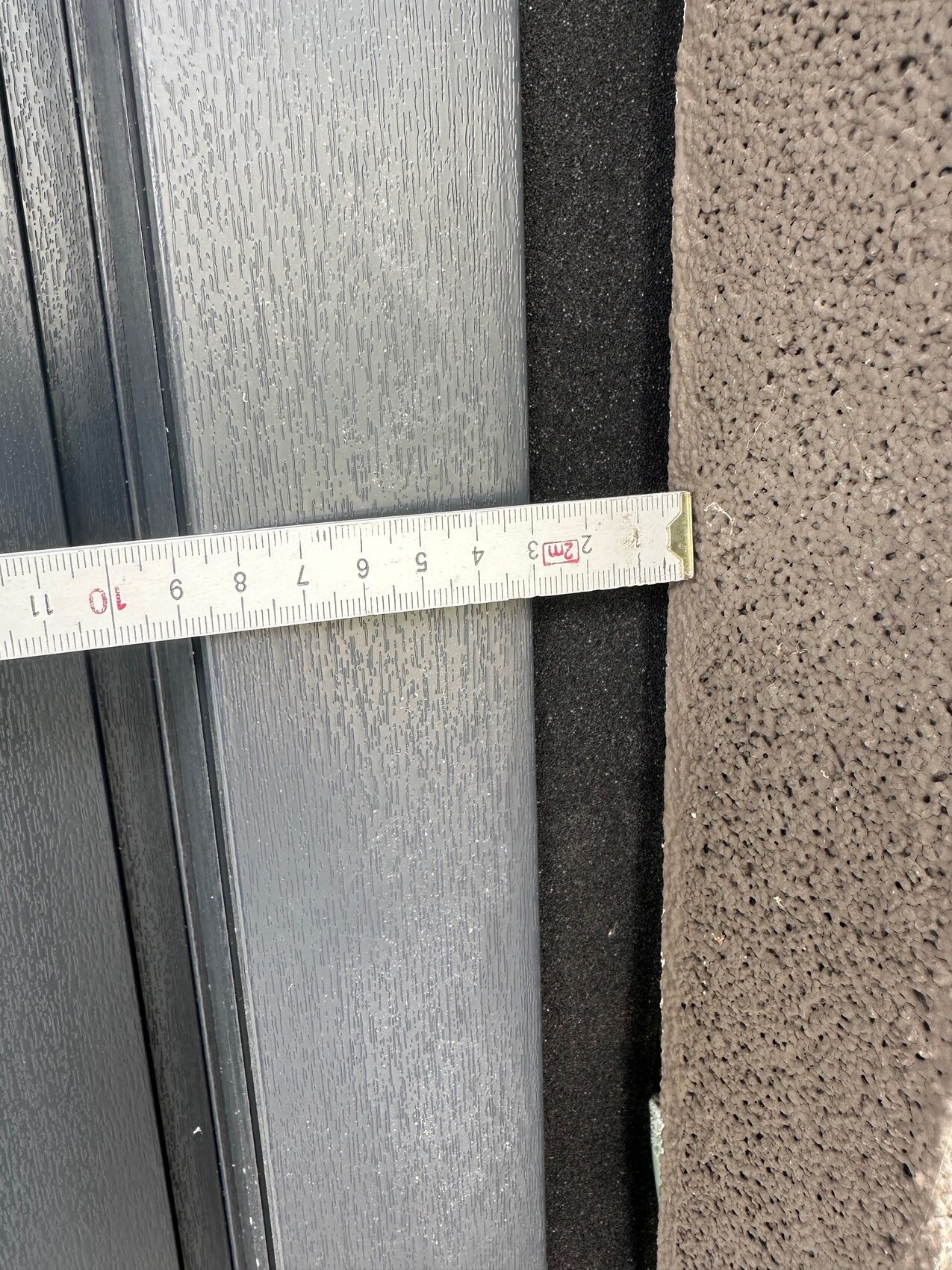 Maßband liegt quer über den Spalt zwischen grauer Türrahmenkante und beigem Teppichboden.