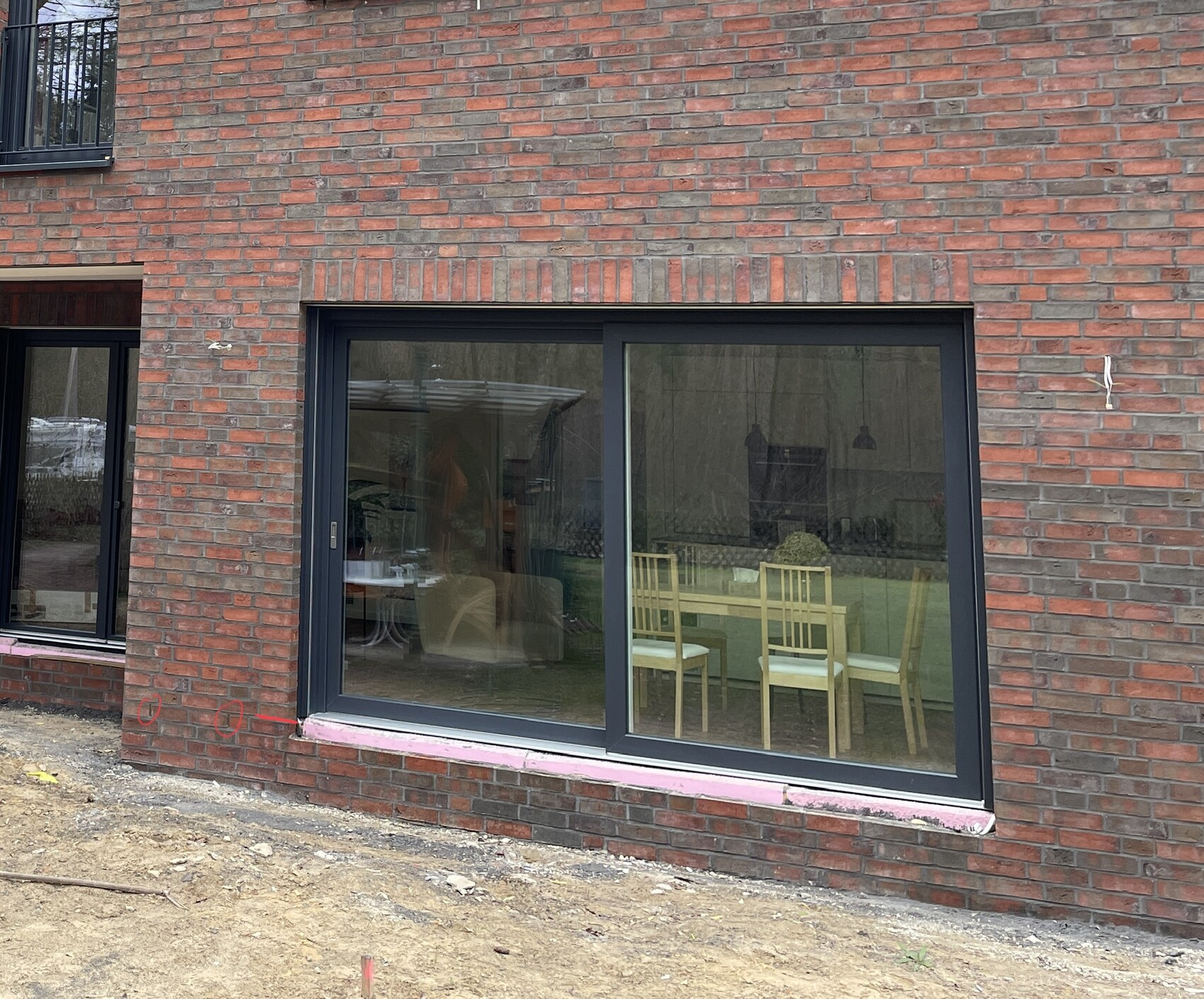 Große Glasfront mit dunklem Rahmen vor rotem Backstein; Einblick in Essbereich mit Stühlen.