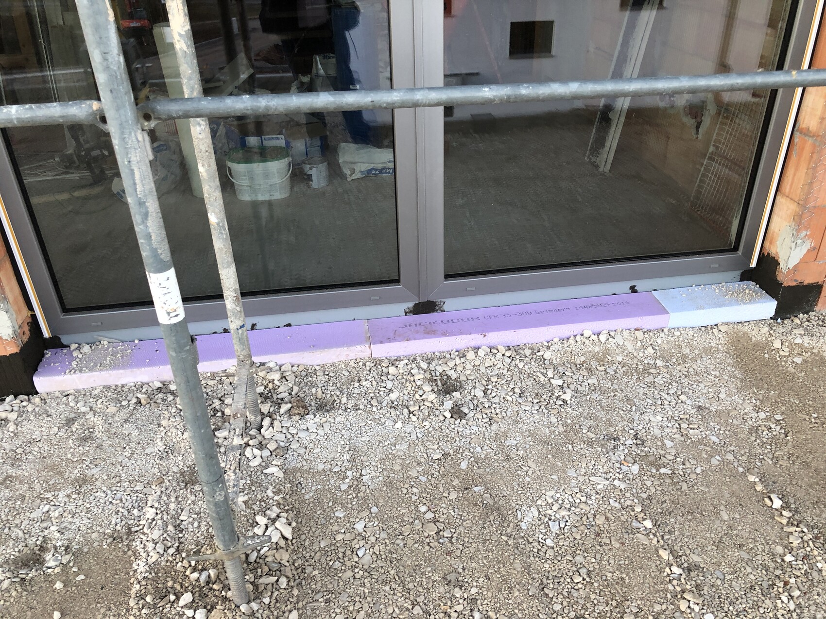 Baustelle: Große Glasfront (Schaufenster) mit Metallrahmen, Gerüststangen davor, Kiesboden.