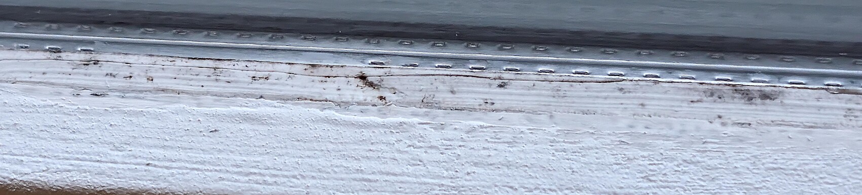 Nahaufnahme eines Fensterrahmens mit Metall-Schiene, rostigen Flecken auf weißer Holzunterlage