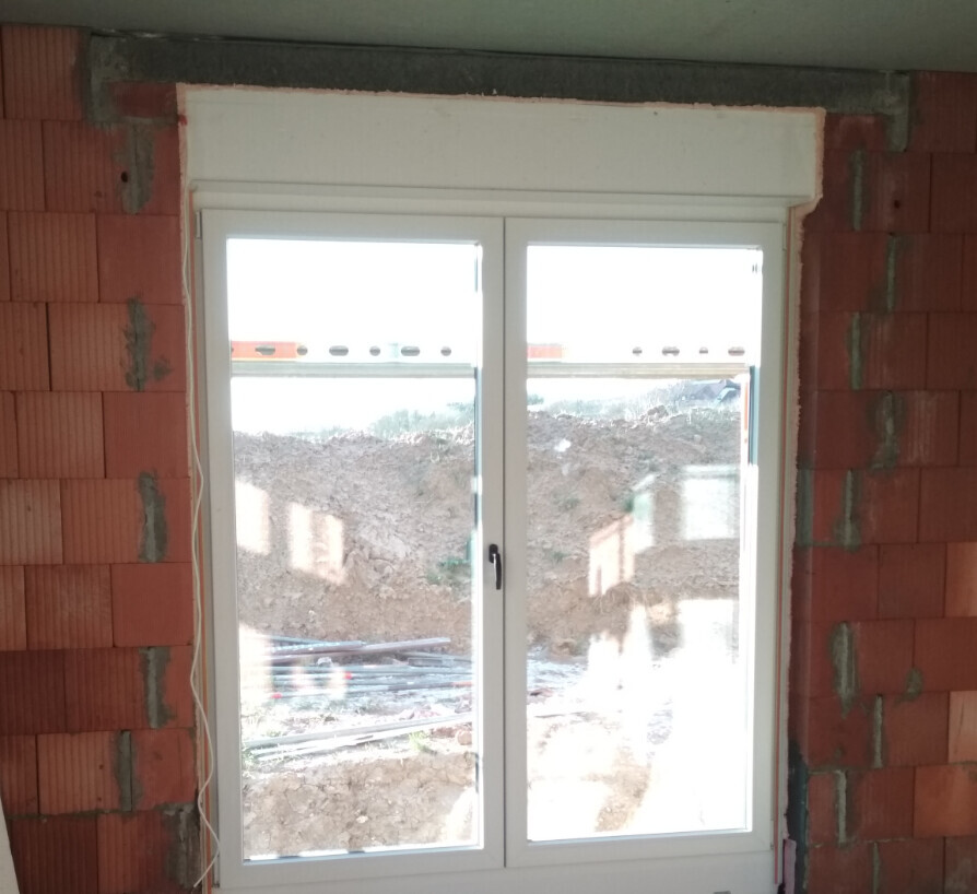 Weißer Fensterrahmen in roher Ziegelwand eines Bauprojekts, Außenbereich mit Erde sichtbar.