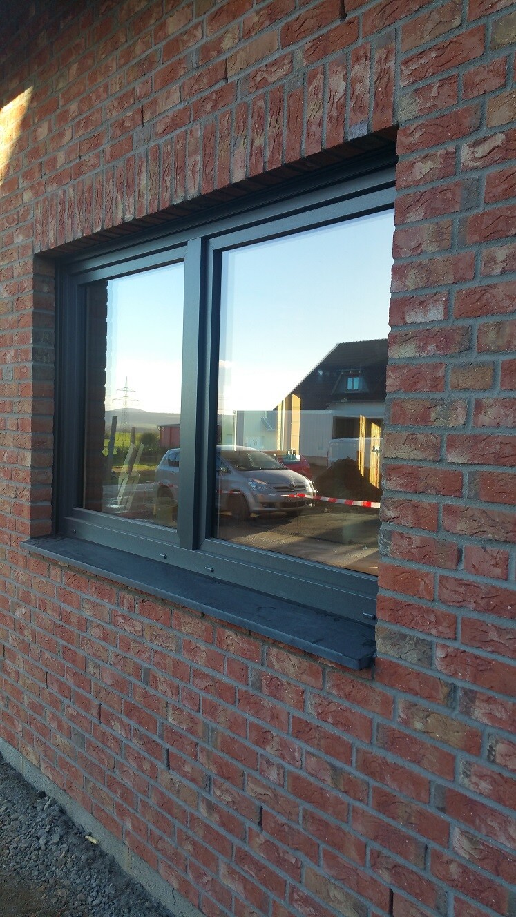 Fenster in roter Ziegelwand mit dunklem Rahmen; Spiegelung eines Autos und angrenzende Häuser.