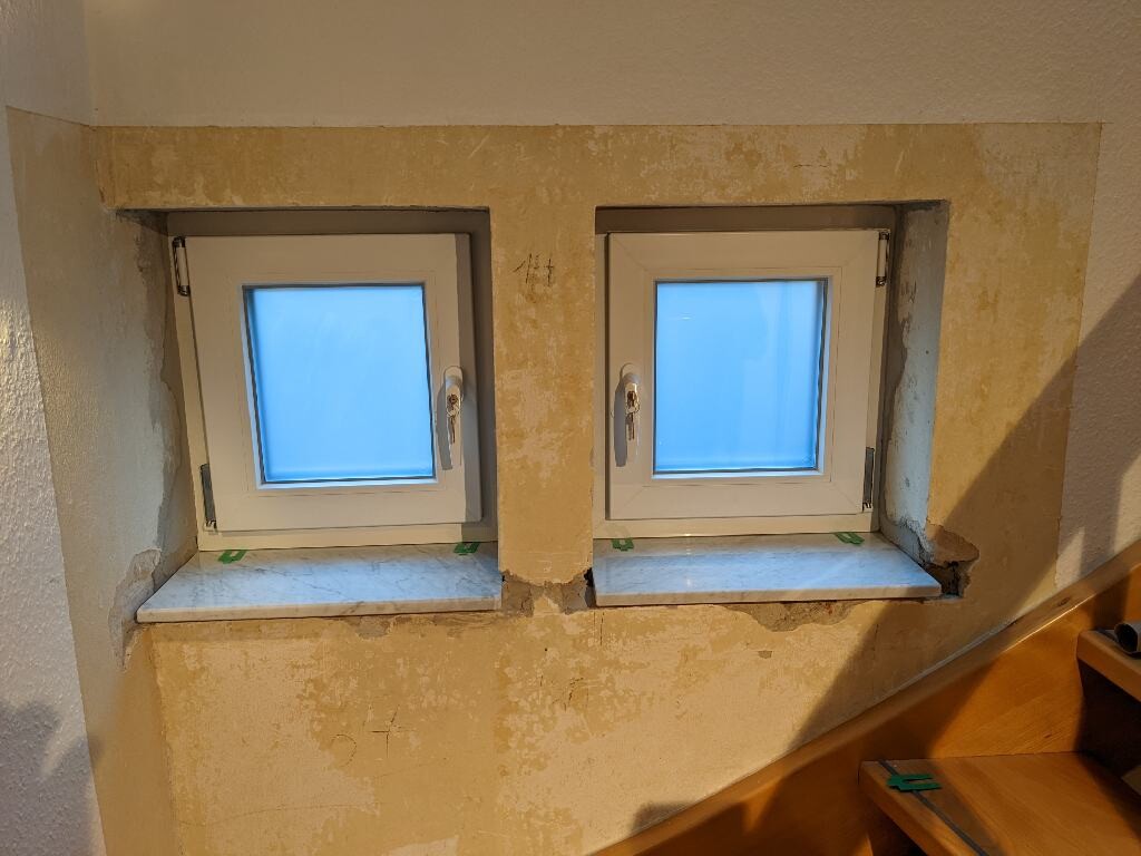 Zwei kleine weiße Fenster nebeneinander in einer gelblichen Wand; rechts eine Holztreppe.