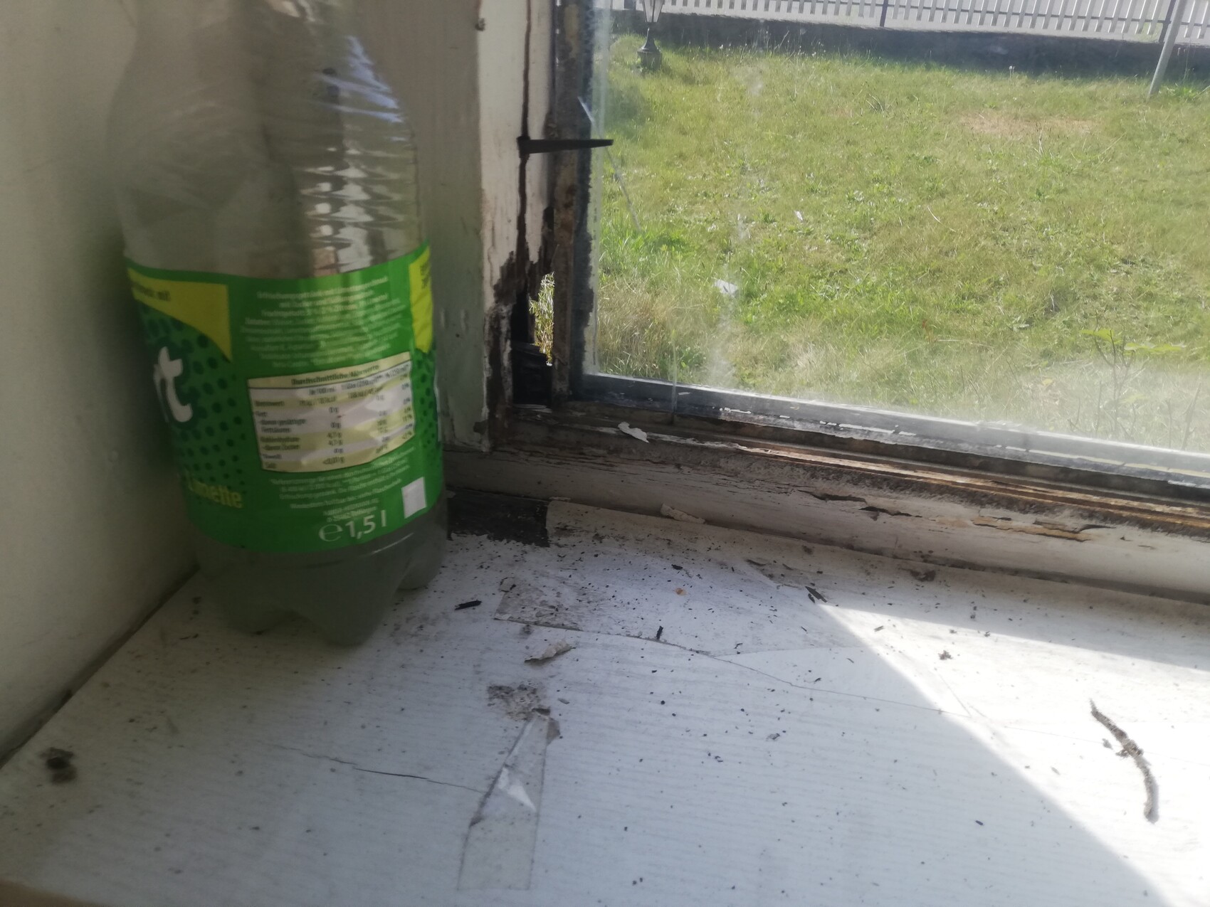 Grüne 1,5-L Flasche neben beschädigtem Fensterrahmen auf schmutziger Fensterbank Gras draußen