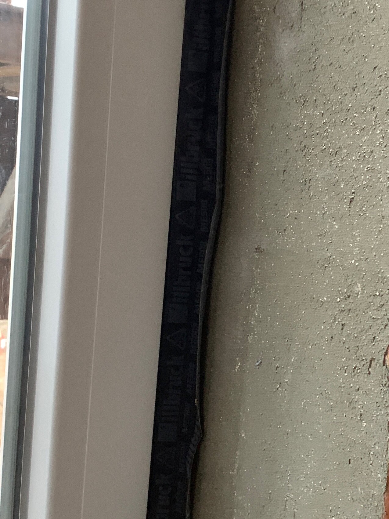 Nahaufnahme eines weißen Fensterrahmens mit schwarzem Dichtungsband neben rauer Wand.