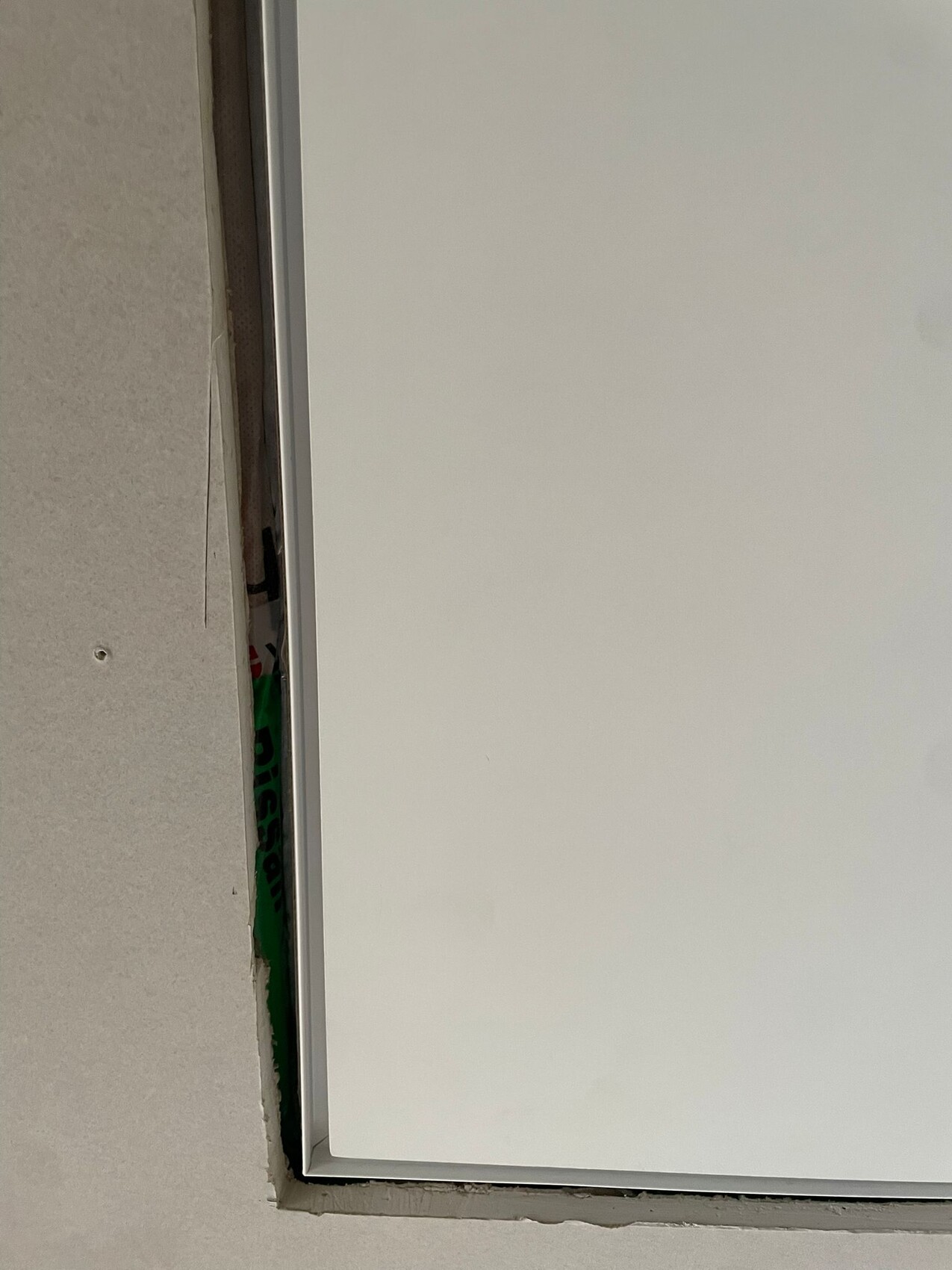 Nahaufnahme eines weißen Fensterrahmens mit Spalt, dahinter grünliches Material sichtbar.