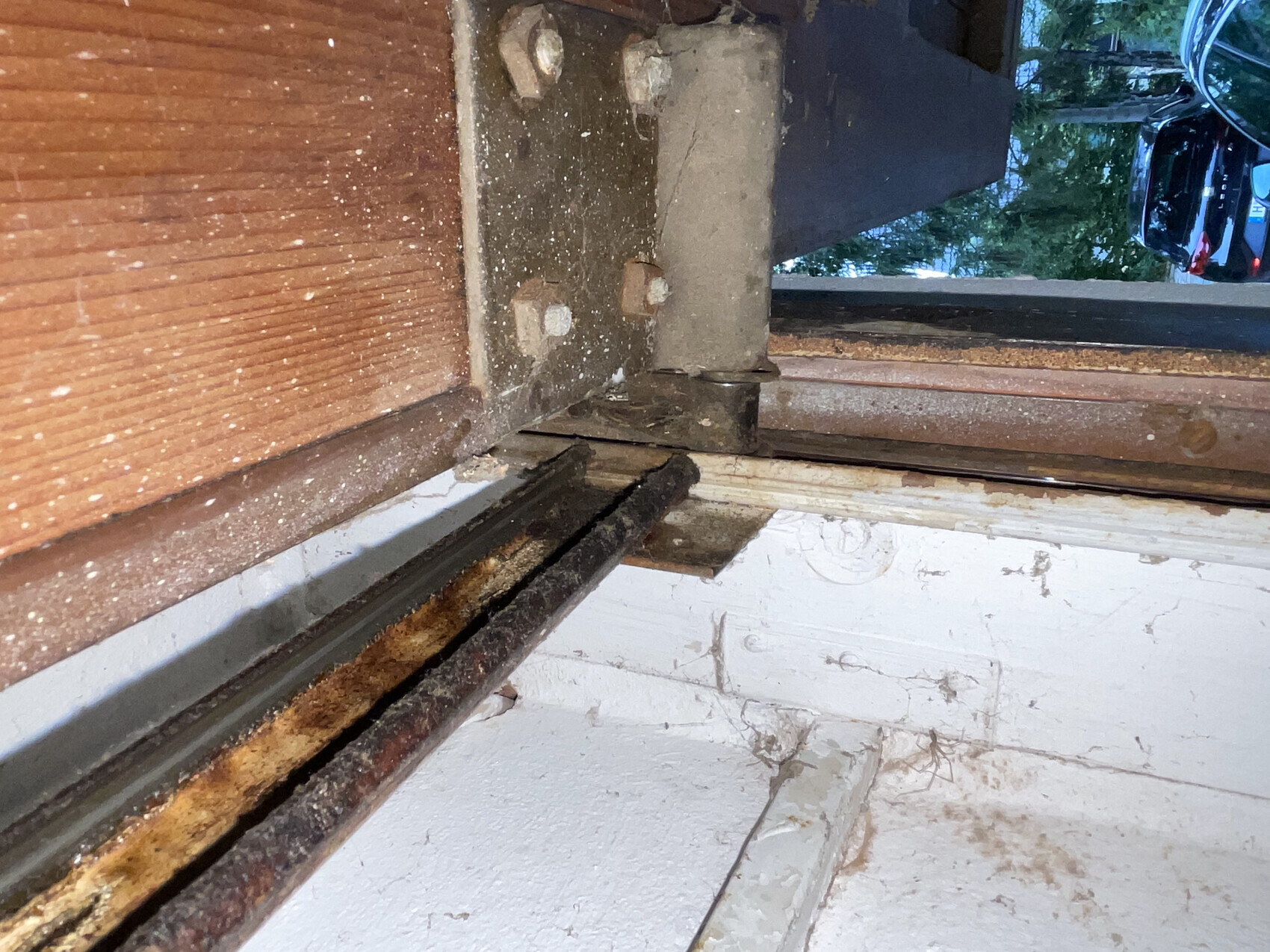 Nahaufnahme einer rostigen Metall-Schiene am Holzfensterrahmen, darunter beschädigte weiße Wand.