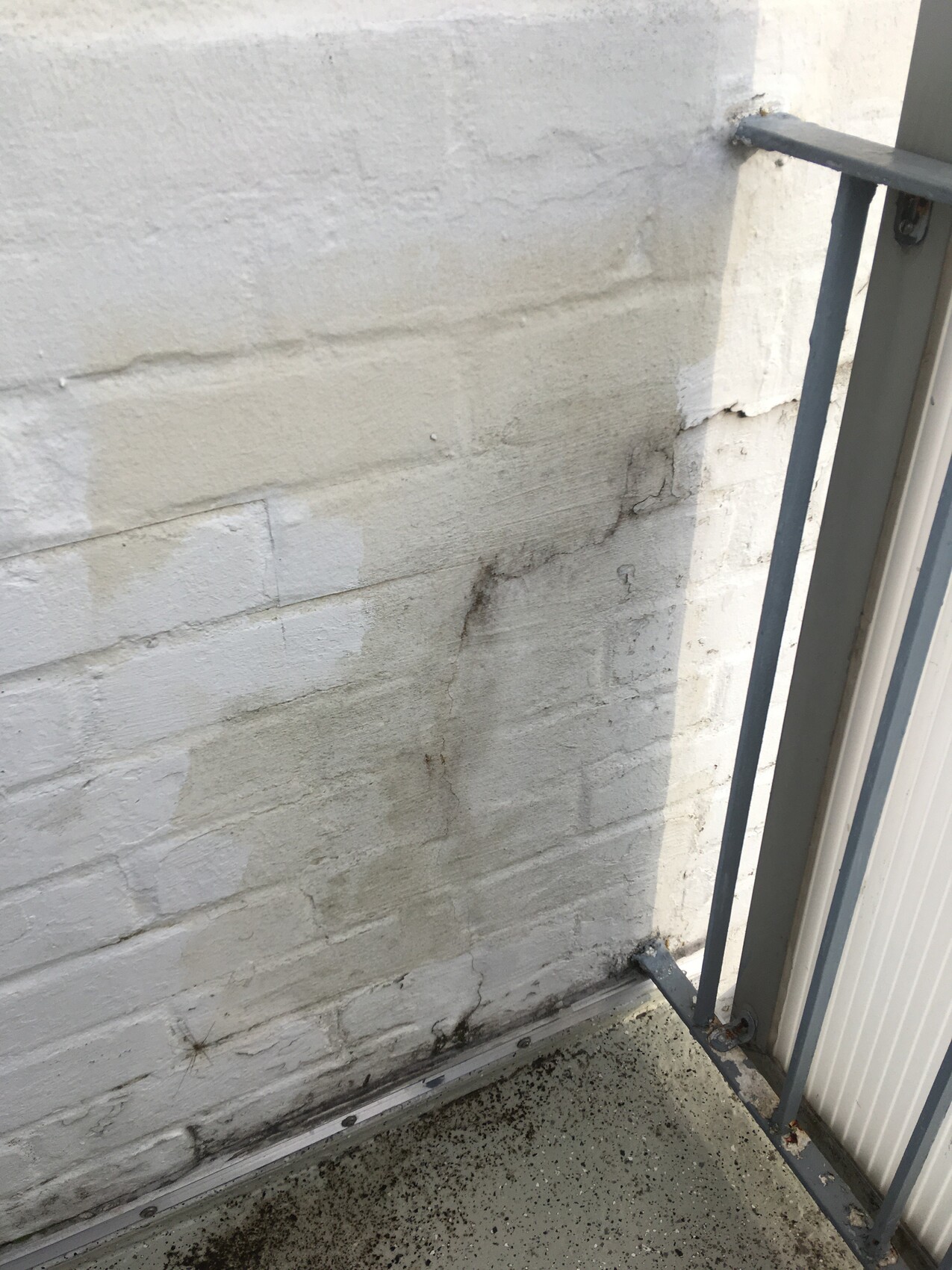 Weiße Ziegelwand mit Feuchtigkeitsspuren und dunkler Verfärbung neben Metallgeländer.