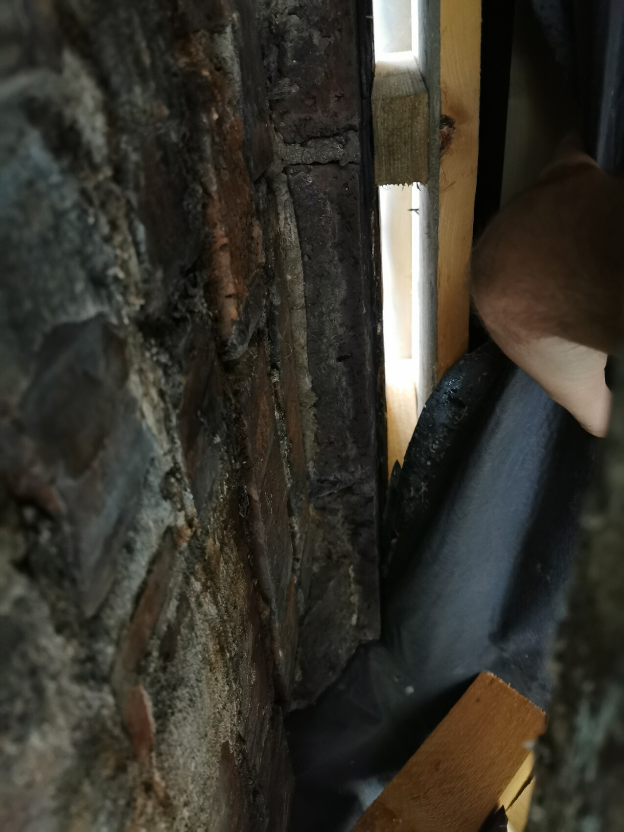 Enger Spalt zwischen Backsteinwand und Holzrahmen, Lichtschlitz sichtbar; Hand und dunkler Stoff davor