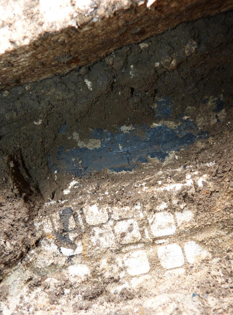 Baugrube mit dunkler Erde, blauer Wandstreifen und hellen Steinpflaster im Boden.