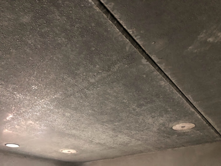 Betondecke mit Feuchtigkeitsspuren, dunklen Flecken und runden Einlässen.