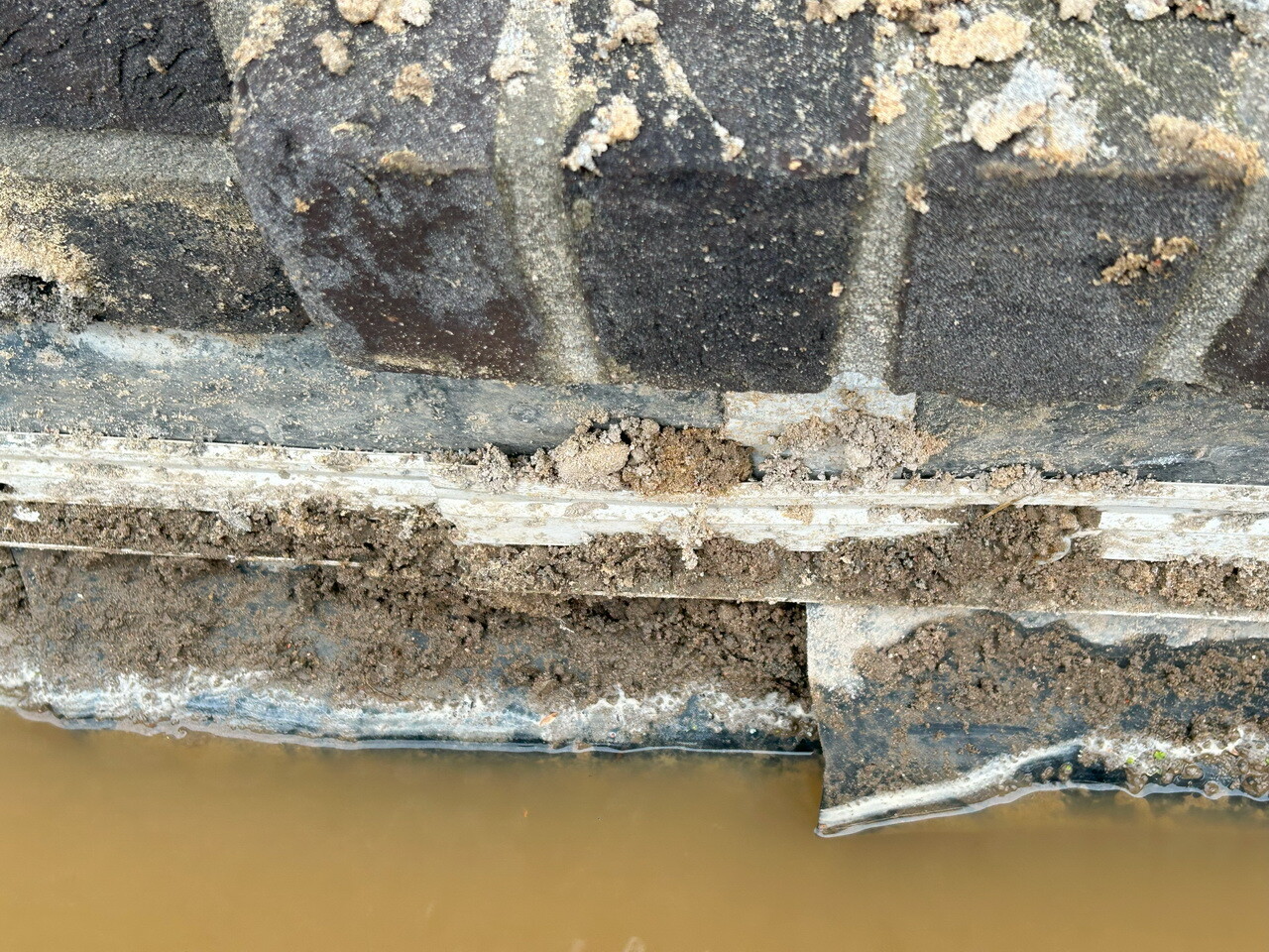 Nahaufnahme einer erdigen Fundamentkante mit Schlamm und braunem Wasser darunter.