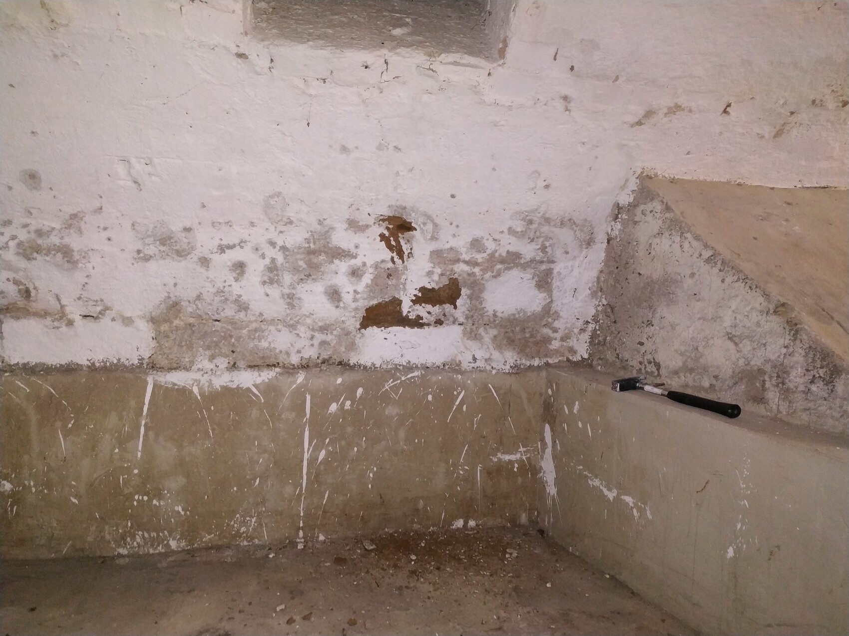 Kellerwand mit abblätterndem Putz in der Ecke; ein Pinsel liegt auf der Kante.