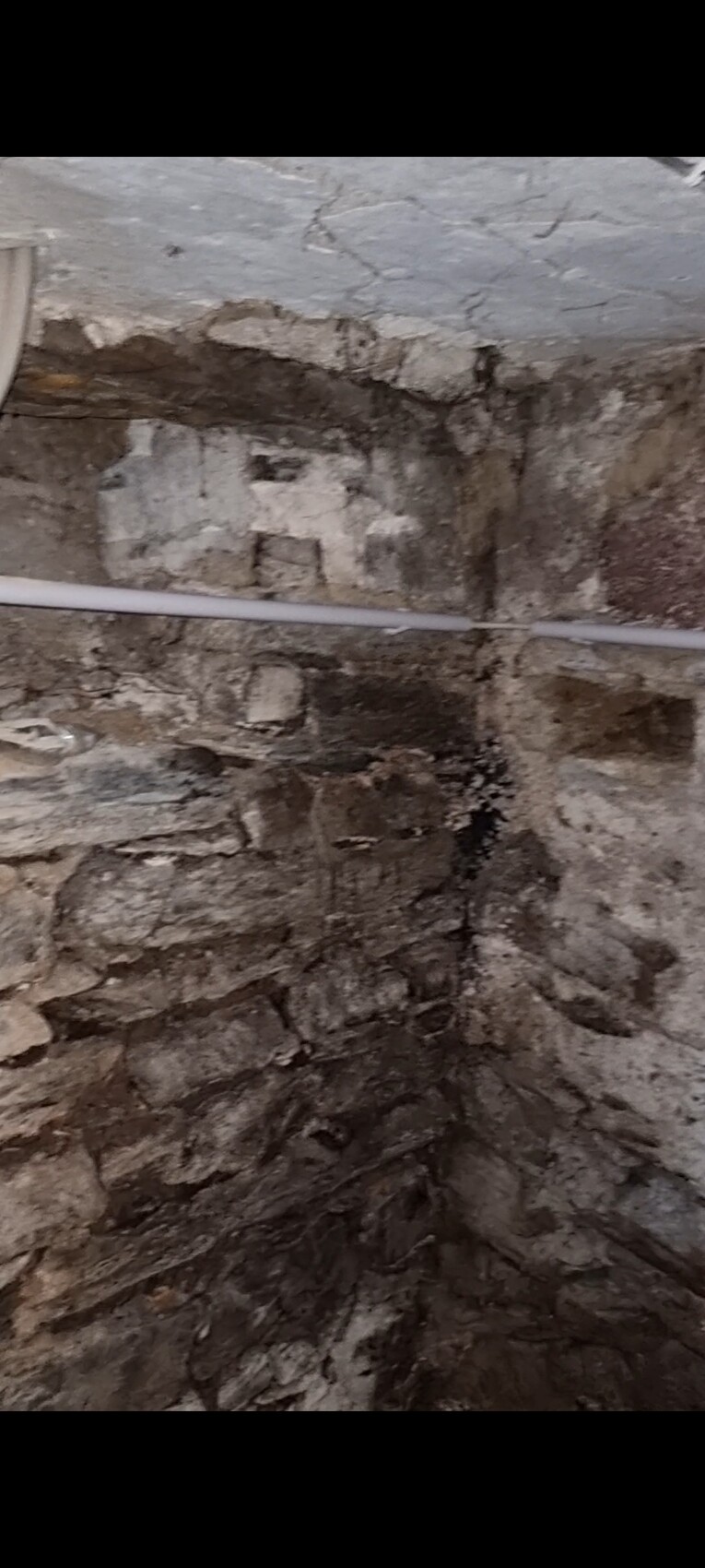 Beschädigte Steinwand mit Rissen in einem Kellerraum, eine weiße Rohrleitung läuft quer hindurch.
