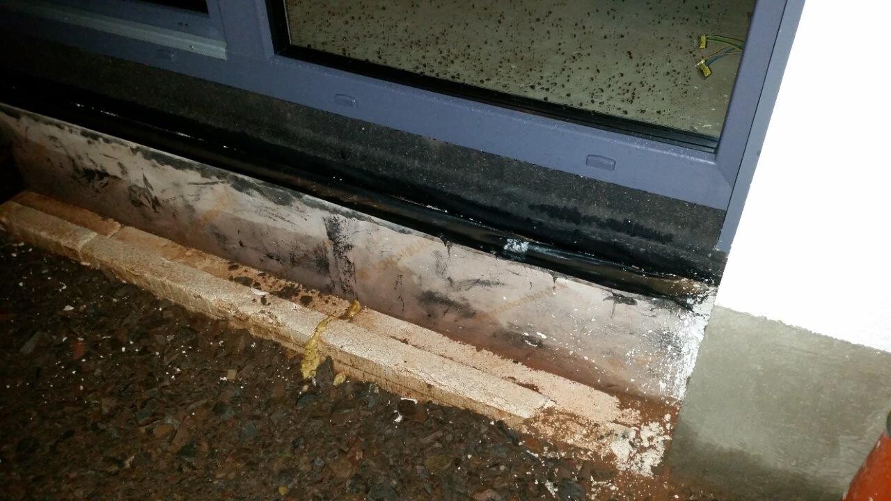 Unterkante eines blaugrauen Fensterrahmens mit feuchter, verschmierter Wand und Schlamm am Eingang.