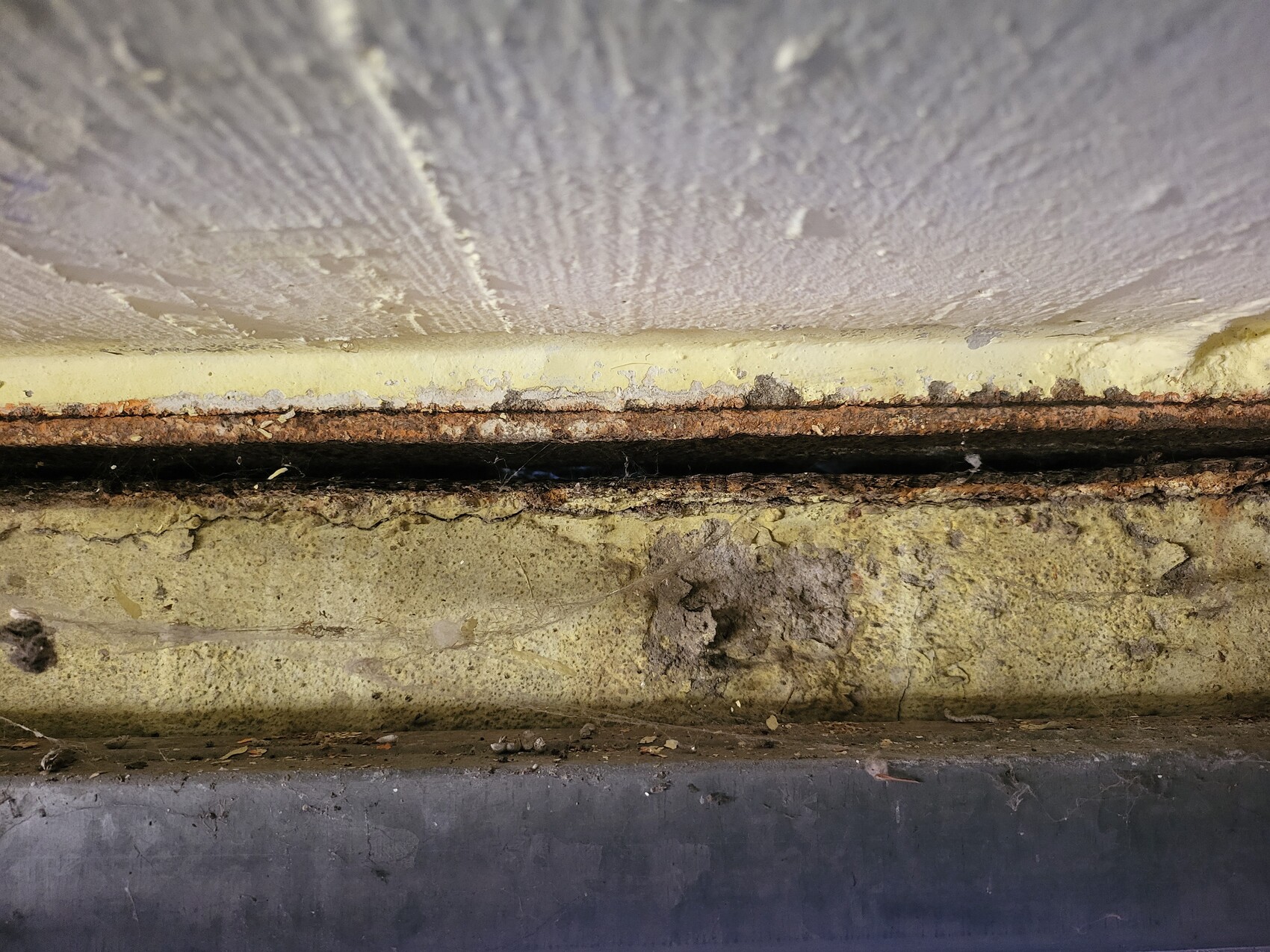 Nahaufnahme eines rostigen Metallstreifens zwischen gelber Wandkante und verdrecktem Betonboden.