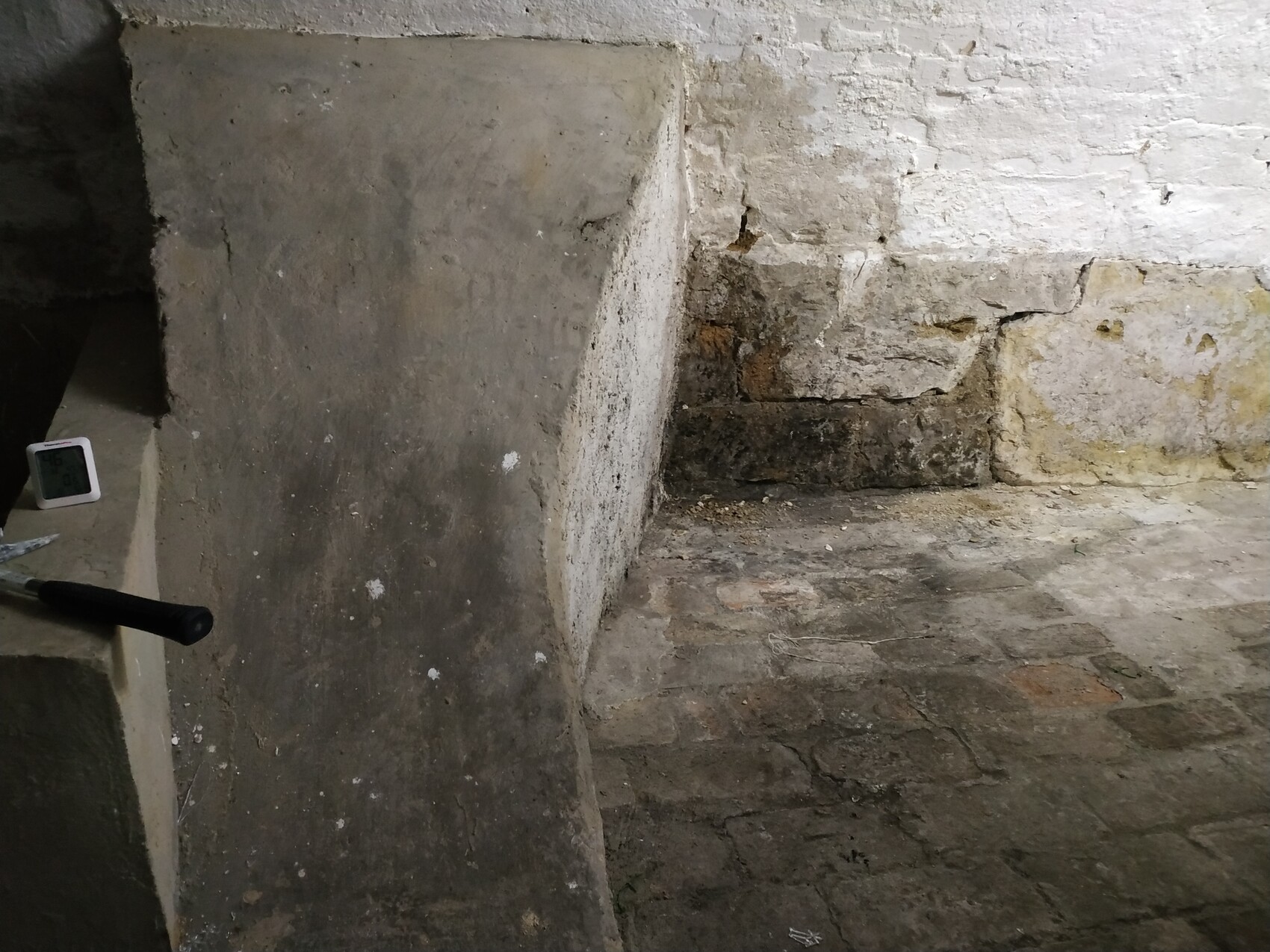 Keller mit weißen Steinwänden und feuchtem Boden; Betonquader; Thermometer und Werkzeuggriff.
