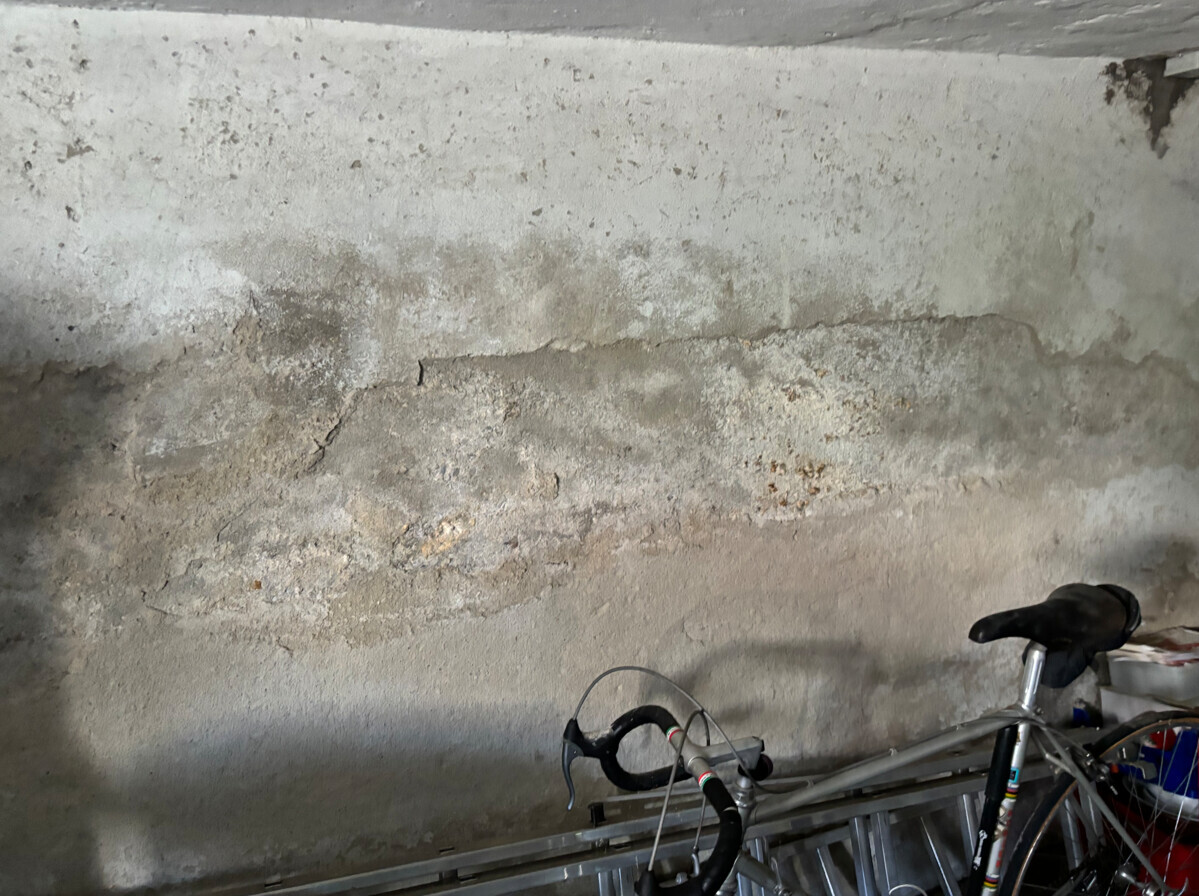 Graue, feuchte Wand mit Rissen und Abplatzungen; im Vordergrund ein Fahrradrahmen mit Lenker.