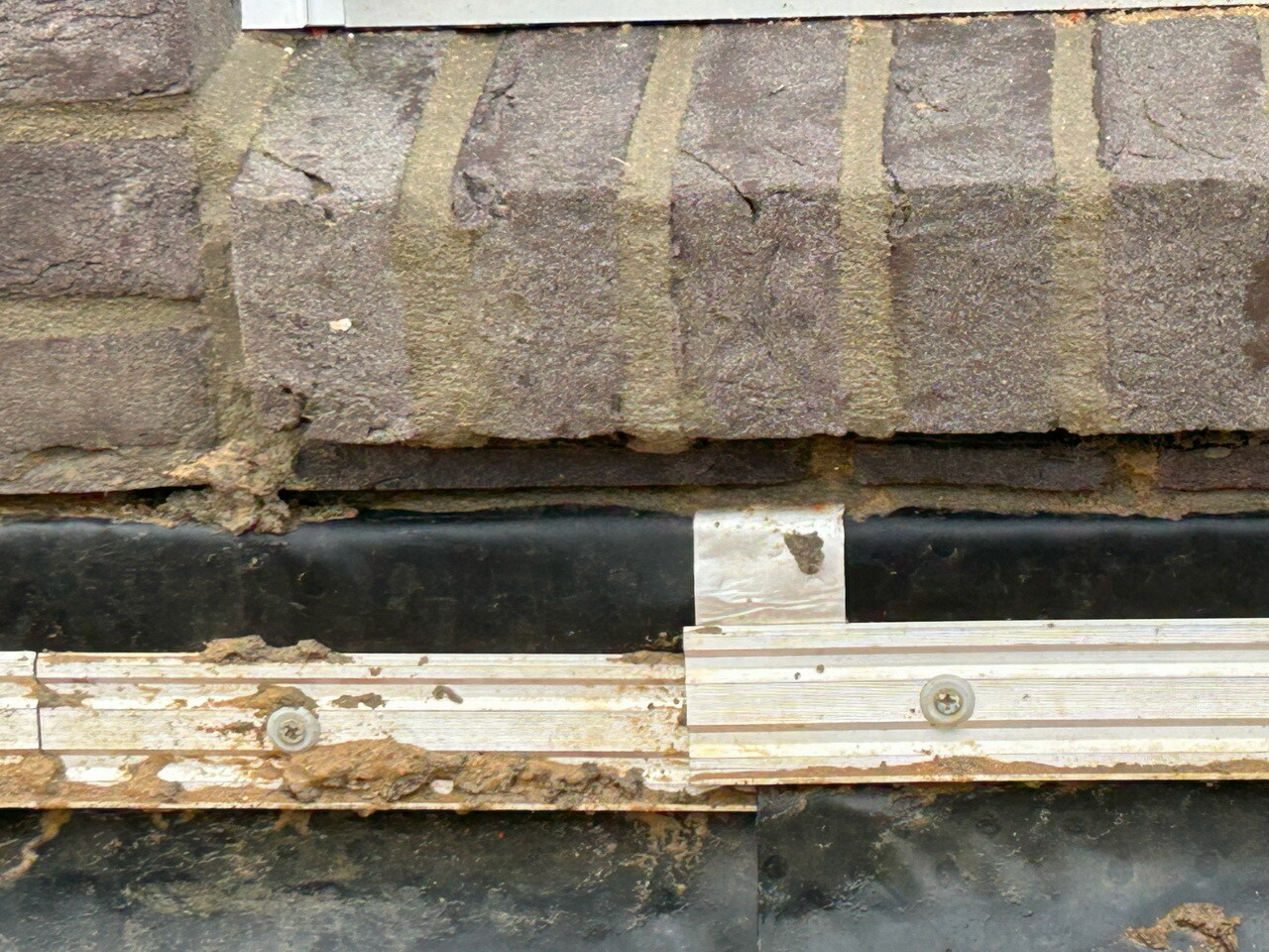 Nahaufnahme einer Steinmauer mit Mörtel, darunter schwarze Folie und eine Holzleiste mit Schrauben.